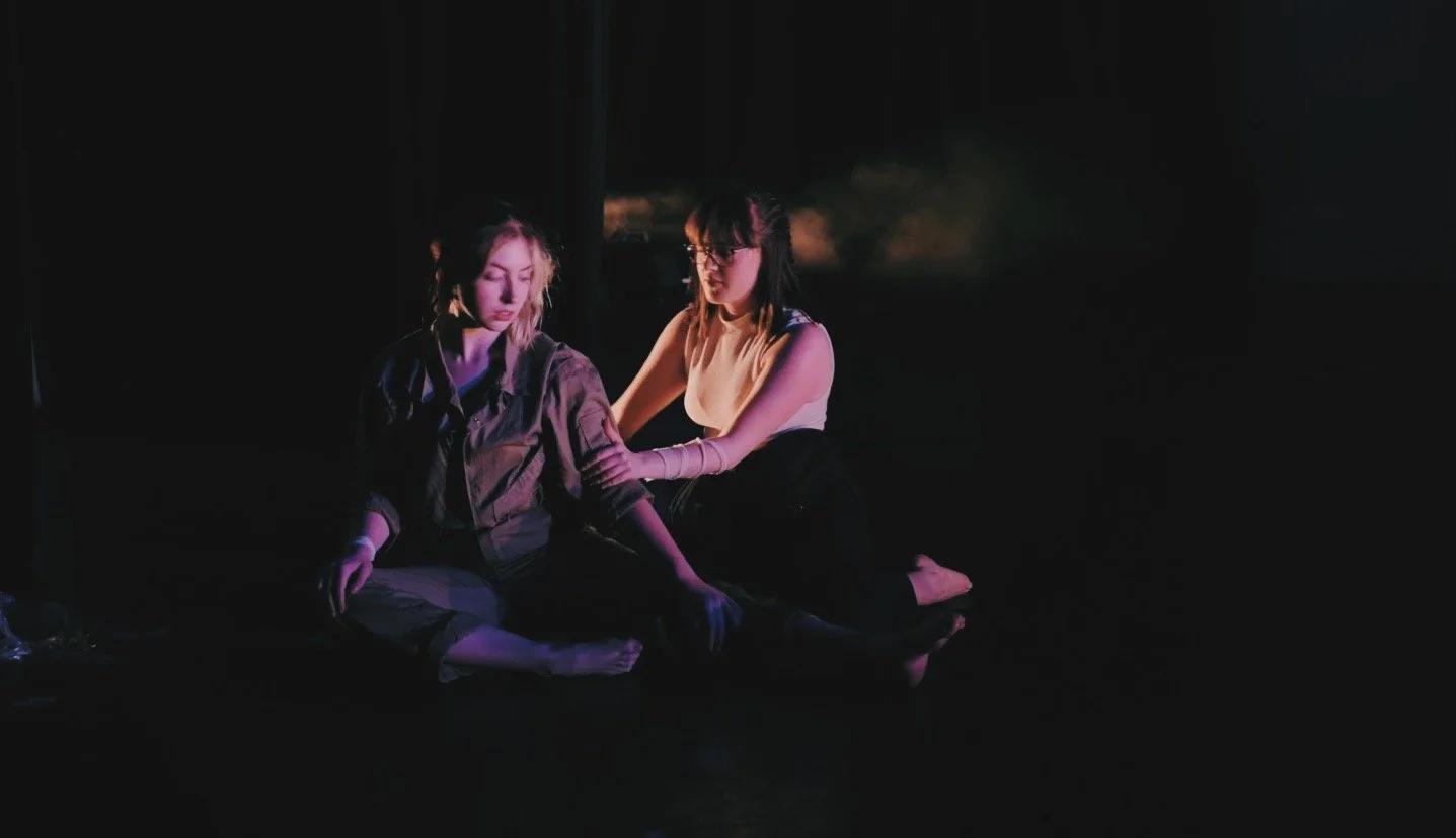 On a very shadowy stage, two dancers sit in fourth, one holding the others arm, both looking tentatively at this touch.