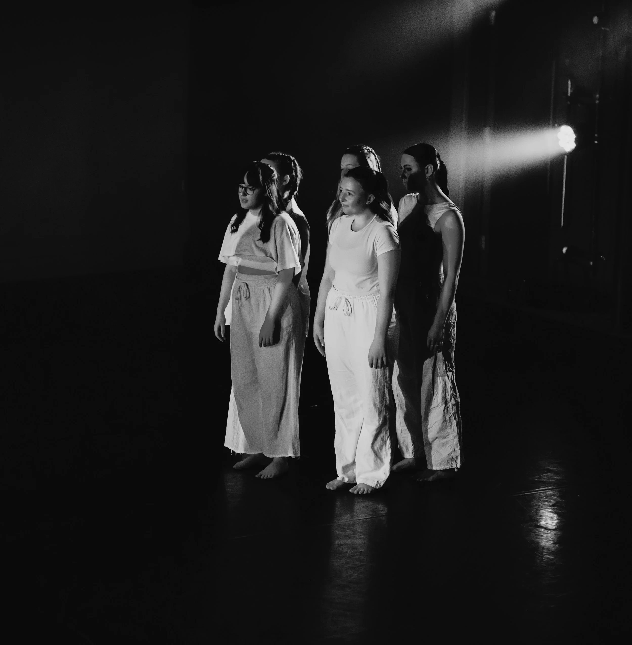 A cluster of five dancers stand on stage lit by warm, red-ish side lights. The are facing stage left and look on adoringly, with a breadth of emotion on their faces.