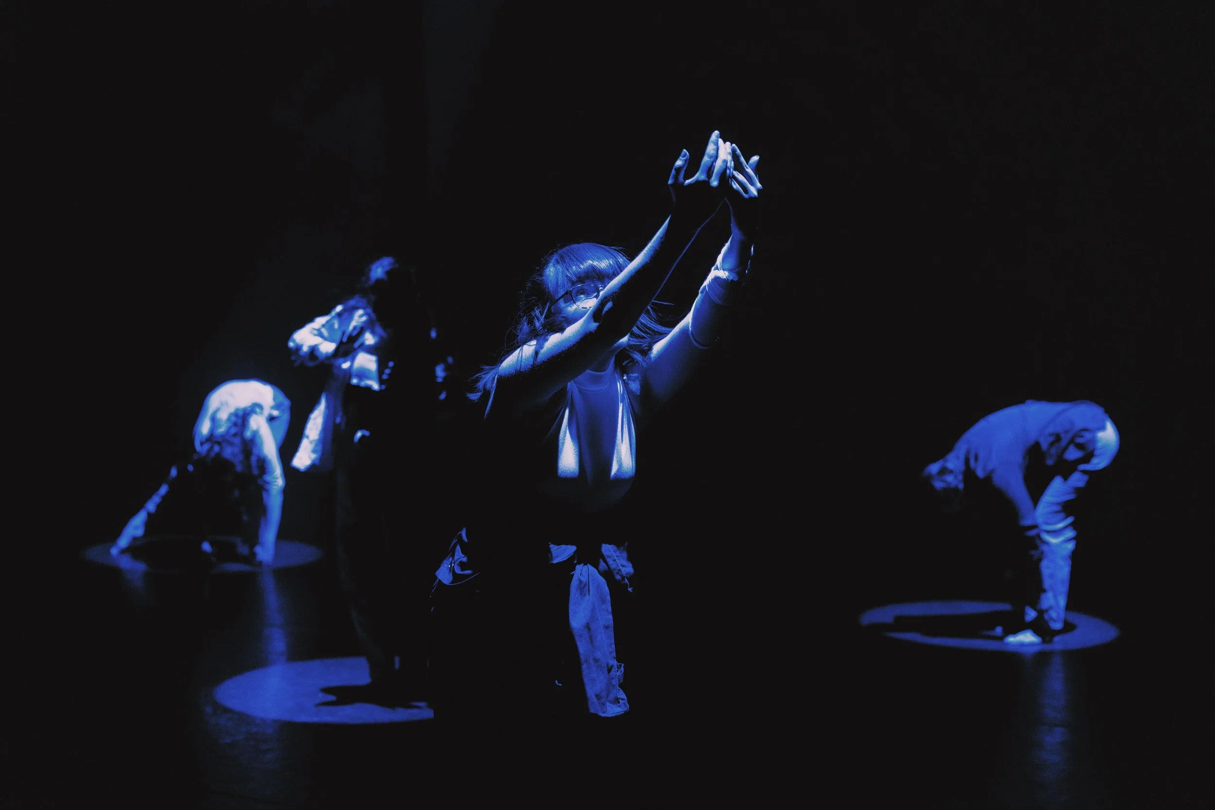 Lit in a bold blue, on a dark stage, a dancer reaches upwards to the right, arms extended, the left one passes across part of her face, hands in a scooping motion. Another dancer is out of focus in another blue pool of light in the background.