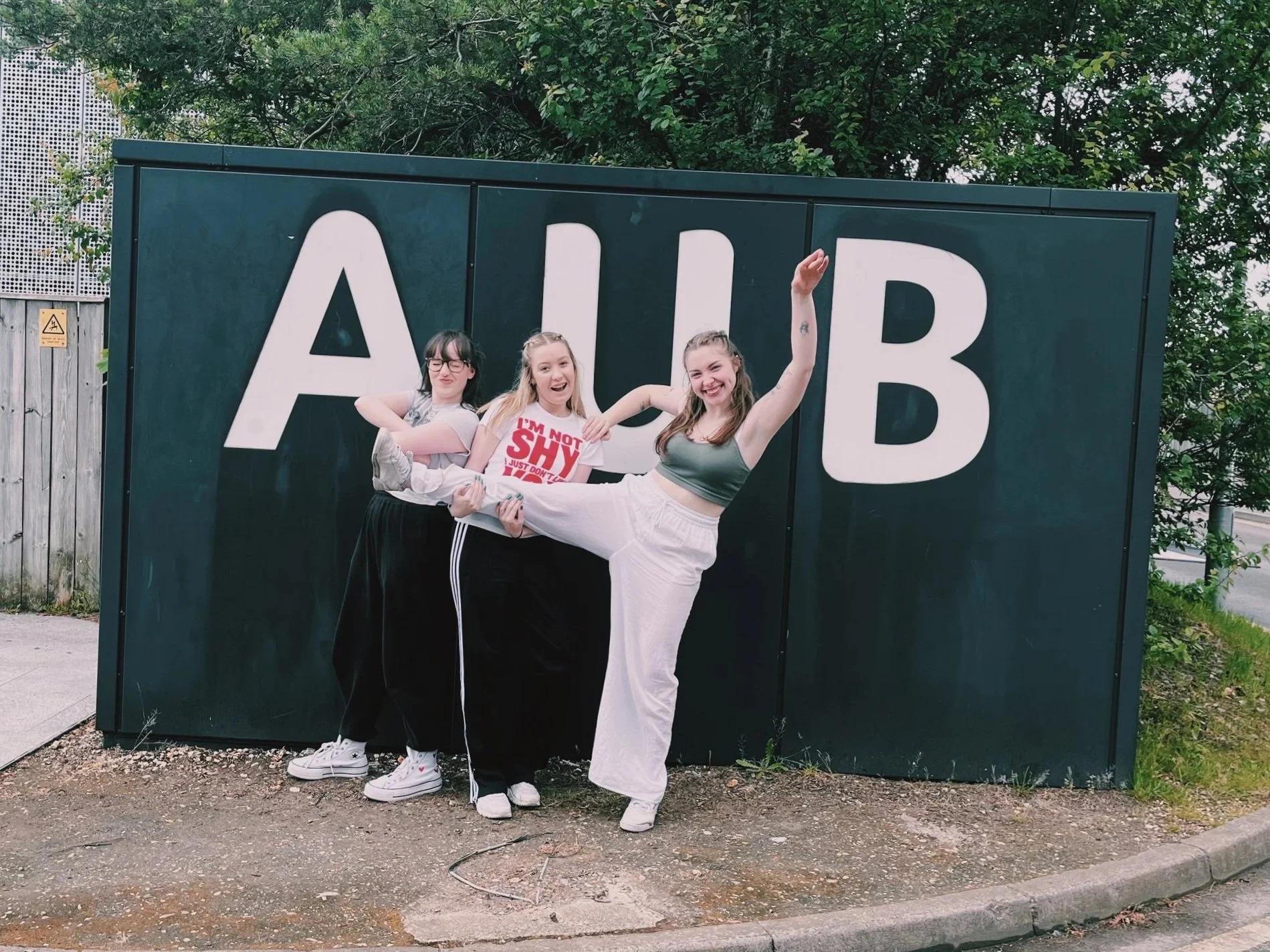 In front of a large AUB sign, three dancers pose for a phot in their training clothes and trainers. On the left on stands arms crossed, back to the middle girl. On the right, the dancer lifts her leg up, held by the middle, outreaching her arms.