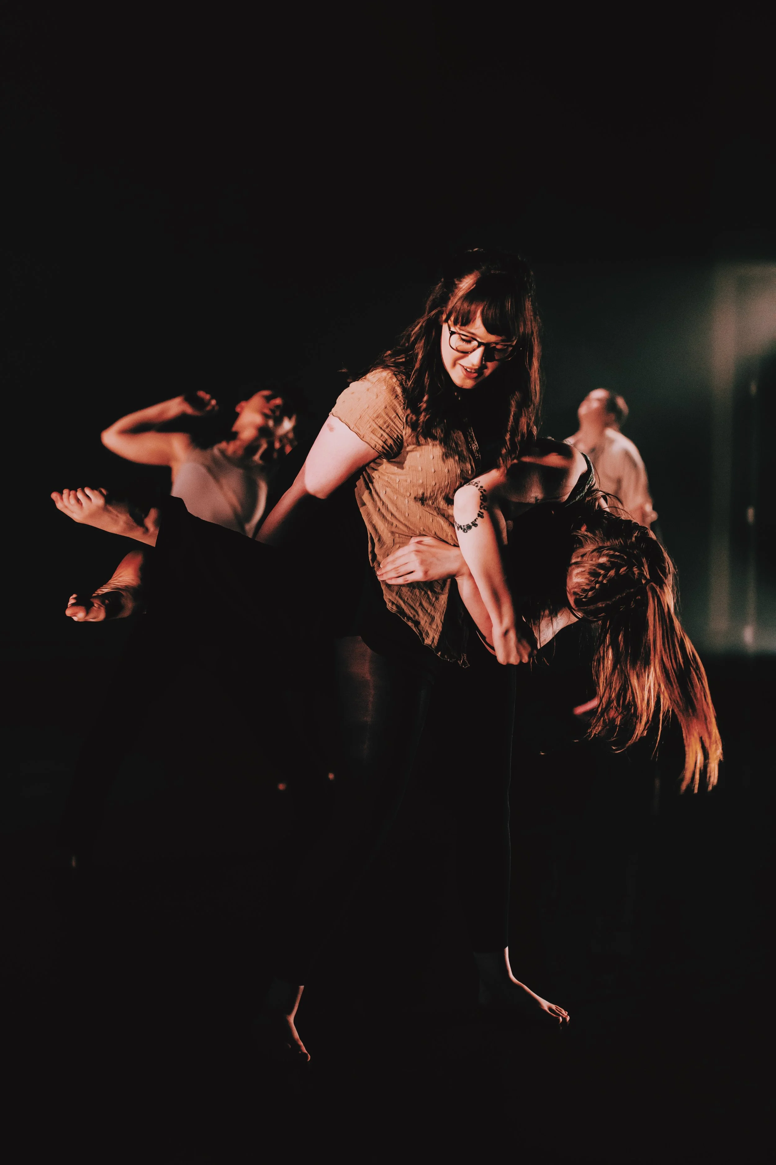 Lit by a warm light on a dark stage, one dancer lifts another. The dancer who is lifted wraps around the others back, cradled in a koala style lift. The base dancer wraps her arms around her and looks down at her tentatively and with care.