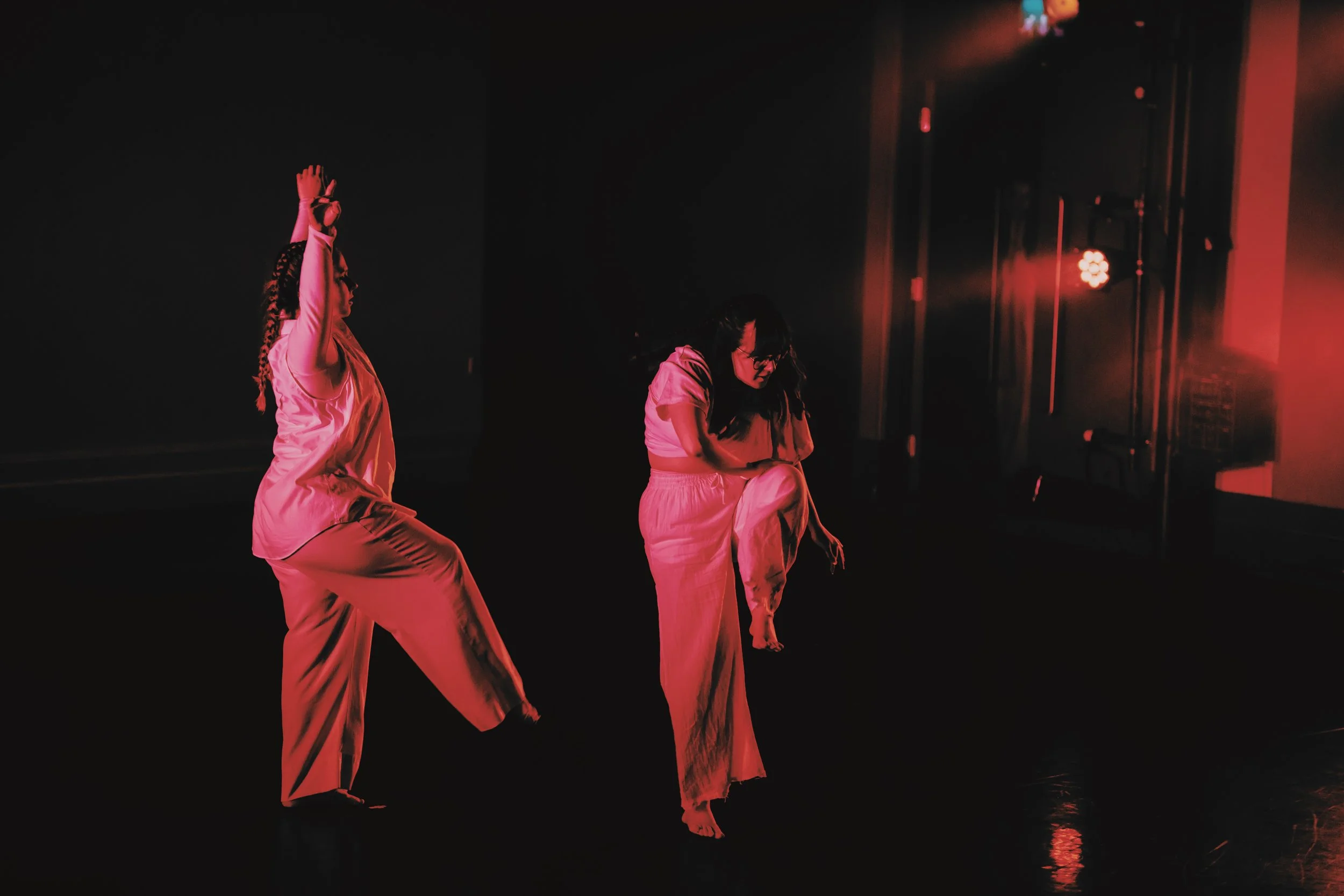 Lit in red, two dancers are in motion on stage. One facing forward, is curved forward, with their knee up, pushing it down away from them. The other faces her, arms upwards reaching outwards above their heads, just beginning to outstretch her leg.