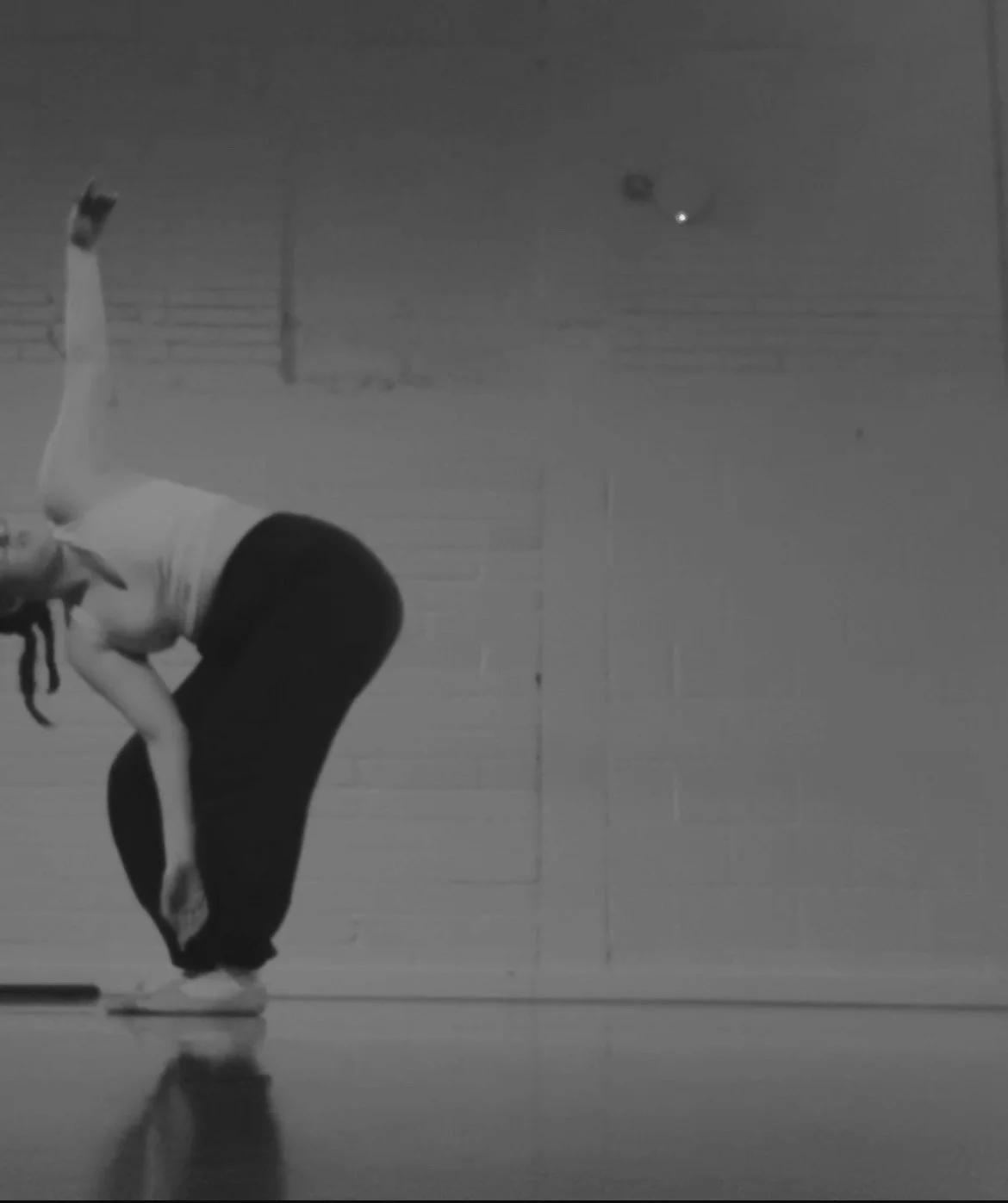 at the edge of the image, a dancer bends down towards the floor, whilst reaching her right arm upwards which opens up her chest and her face looks up to the arm above.