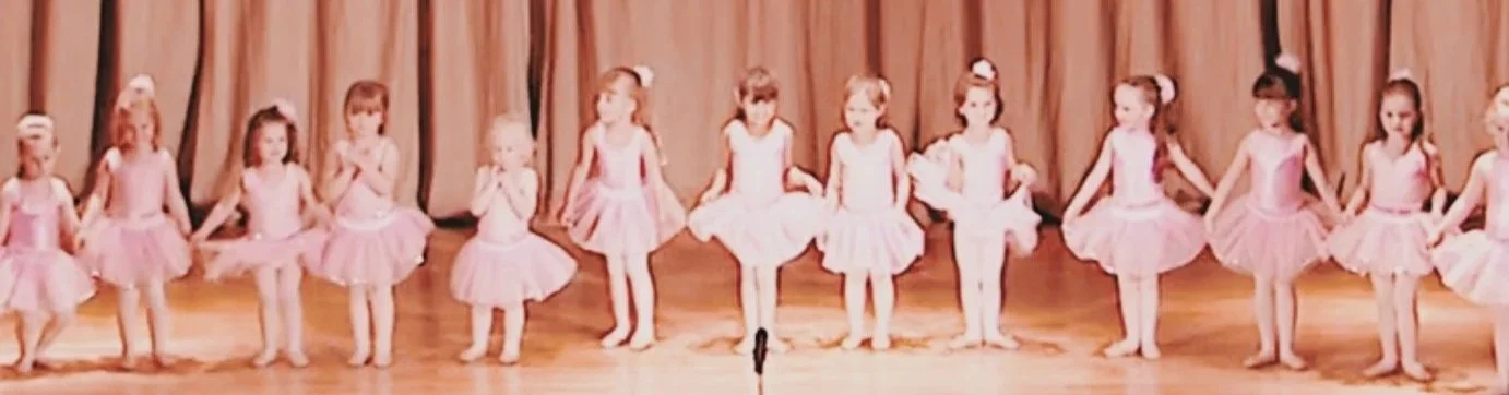 12 baby ballerinas line up on stage. They're holding their tutus and stood in first position. All of them are dressed all in pink with a pink scrunchie in their hair.