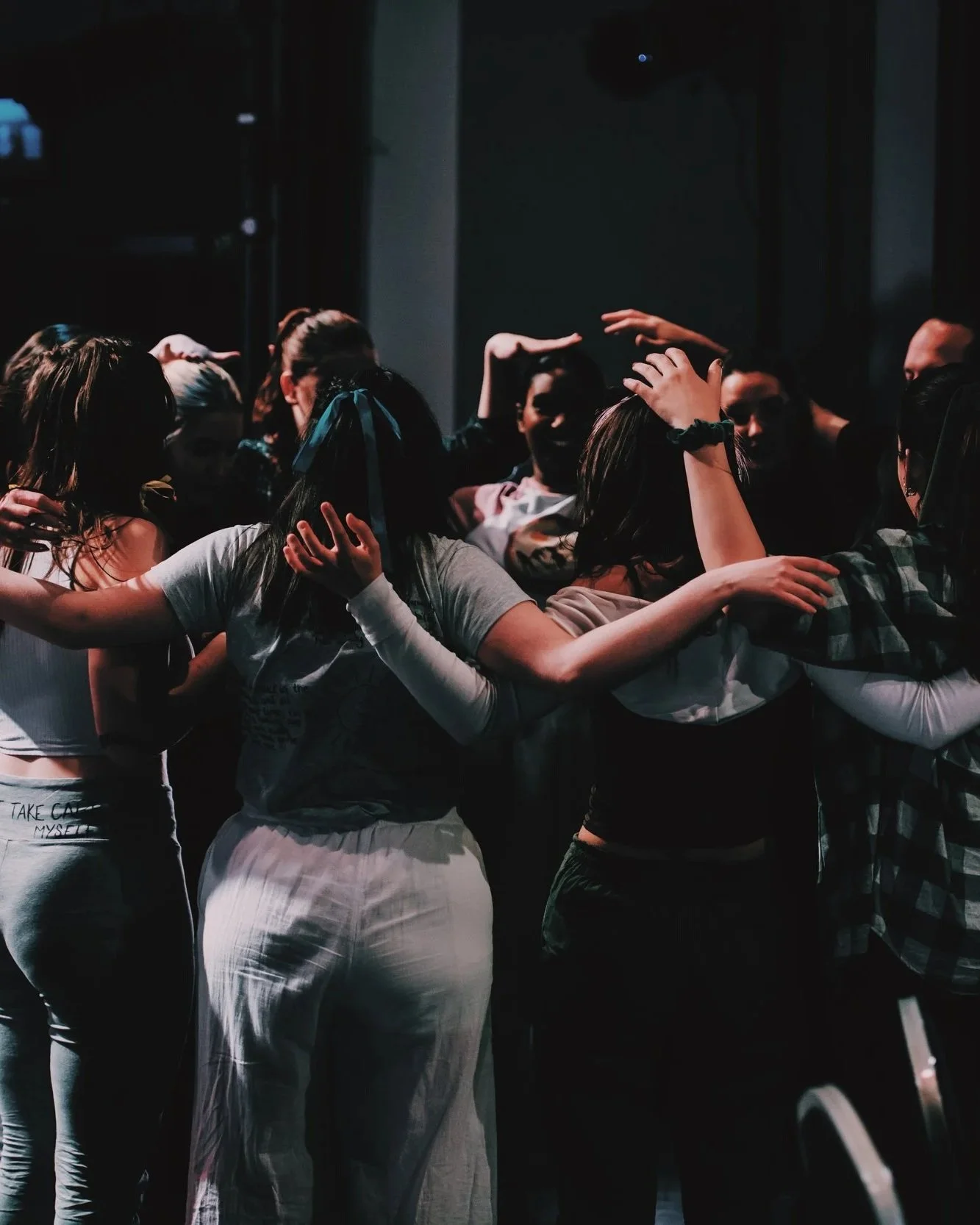 A big group of dancers stand in a huddle, arms all wrapped around eachother, some petting eachothers head adoringly, in a group warm up on the stage pre-show.