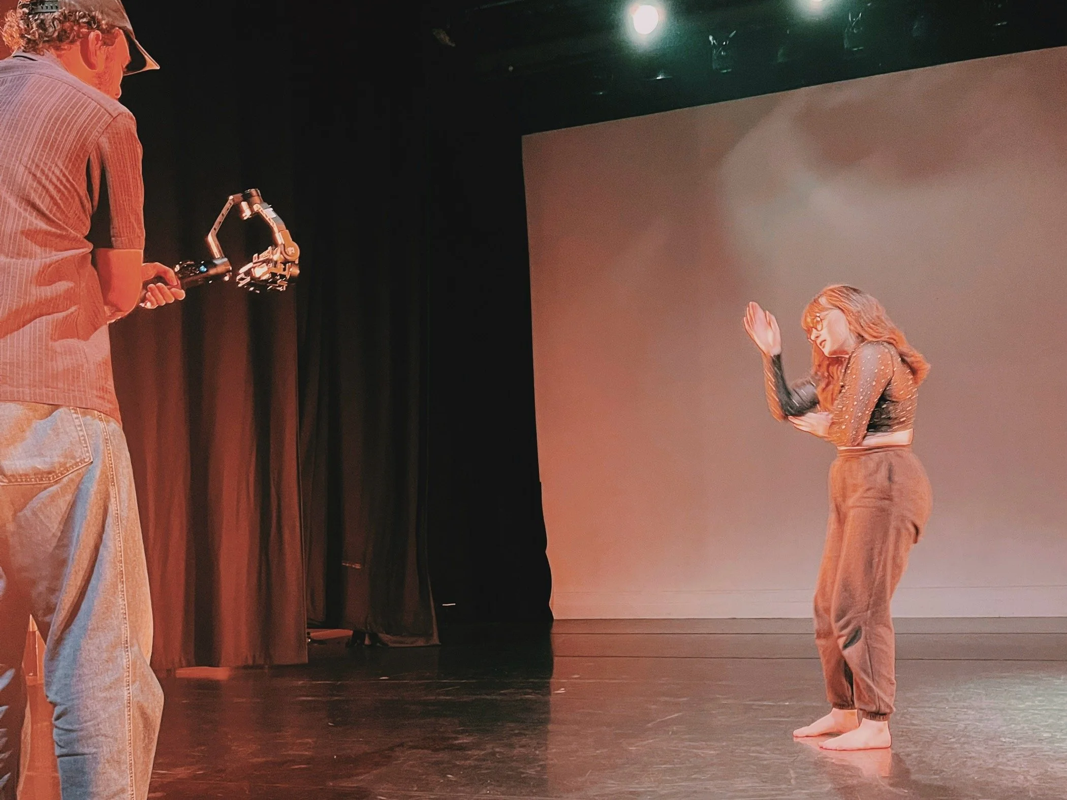 A camera man with a mobile rig stands close to the left side of the image filming the dancer lit by an orange glow. The dancer 's right arm is palm up near her face pulling downwards which will have a knock on effect to move her body backwards.