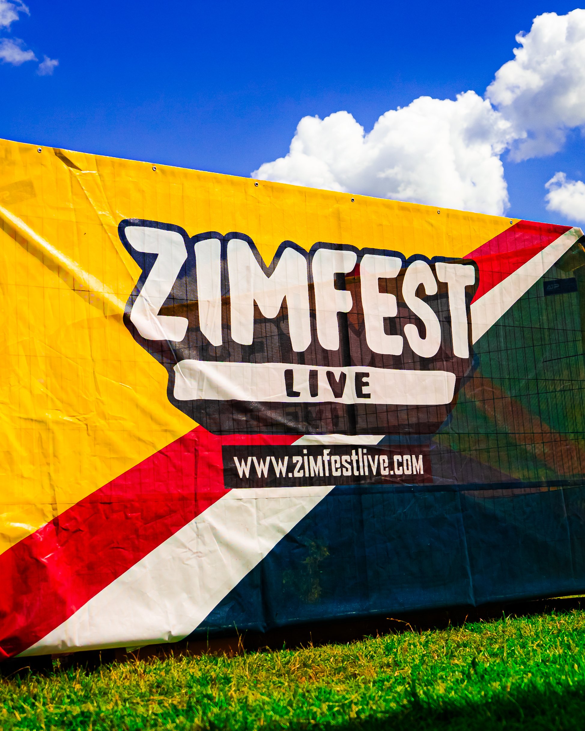 Colorful fabric banner for Zimfest Live festival, with the website www.zimfestlive.com, set outdoors against a blue sky with clouds and green grass.