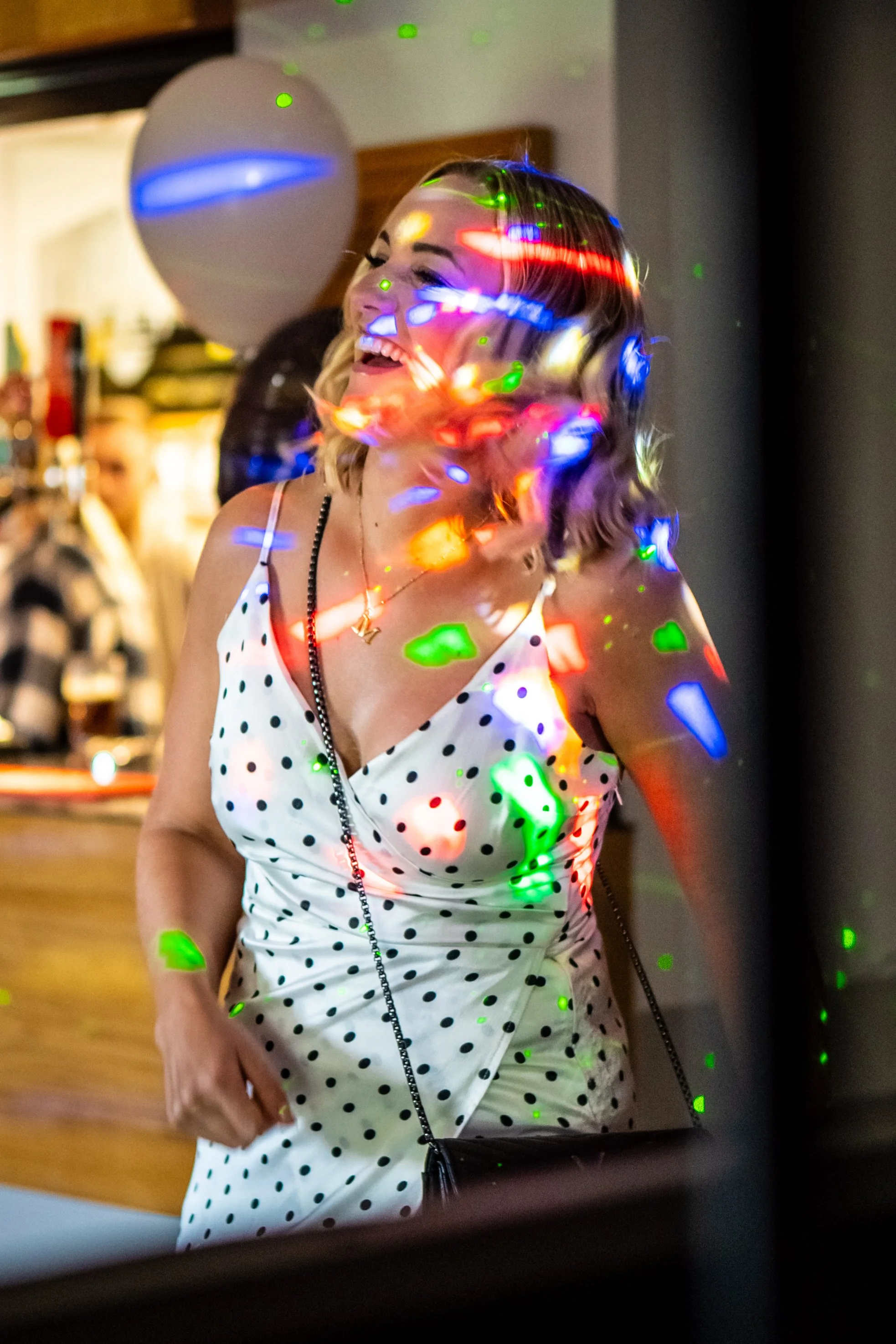 A woman in a white polka-dot dress with a small purse is smiling at a party, with colorful laser lights projected on her face and clothing.