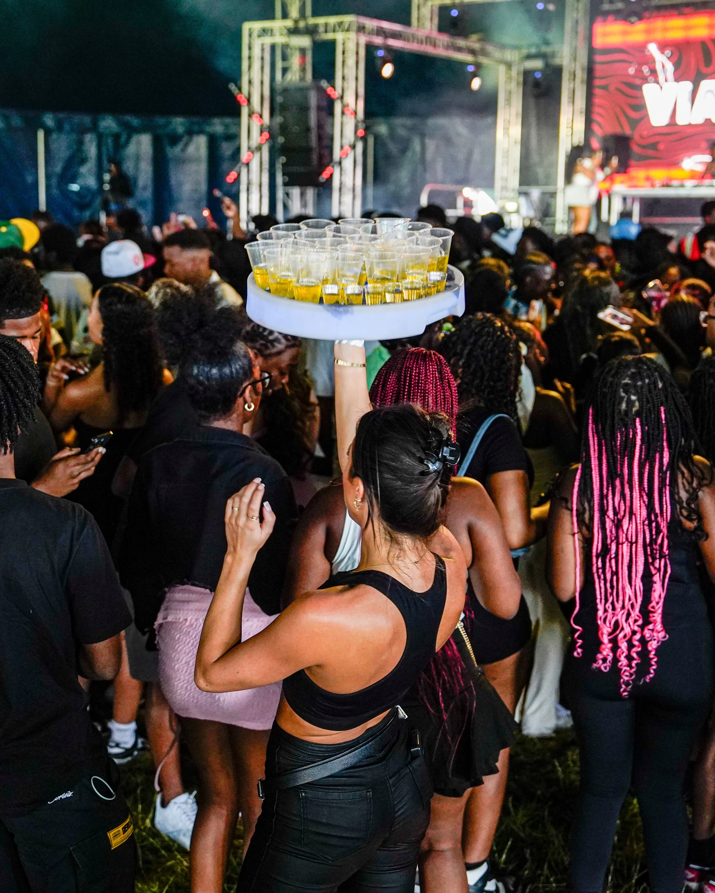 A woman in black clothing holds a large tray with cups of yellow drink at a crowded outdoor event or concert.