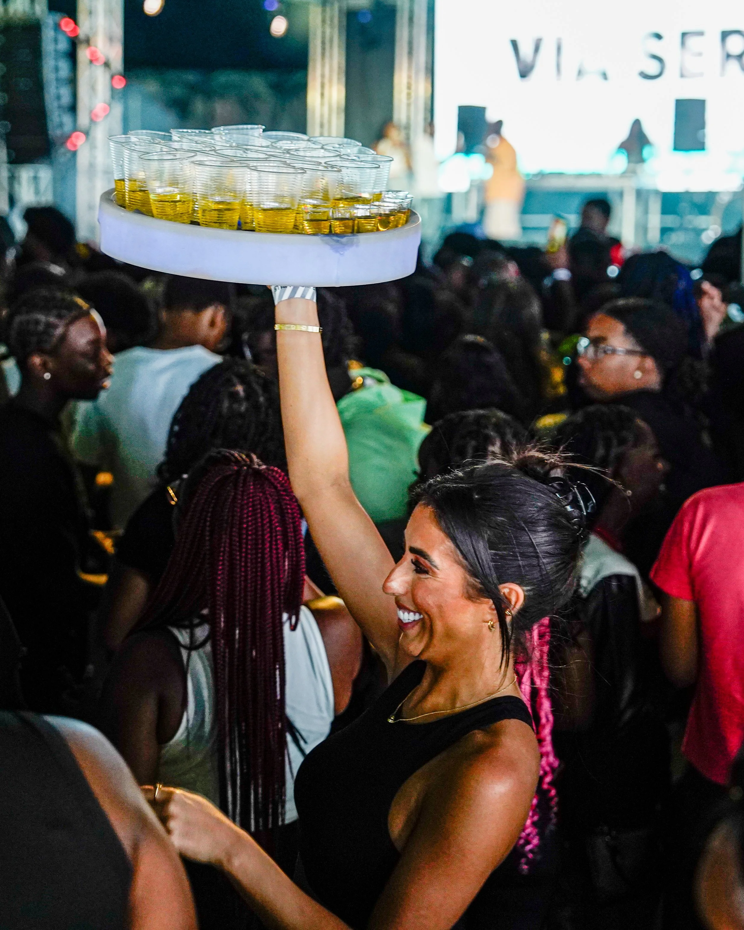 A woman smiling and holding a tray of drinks in a crowded party or event.