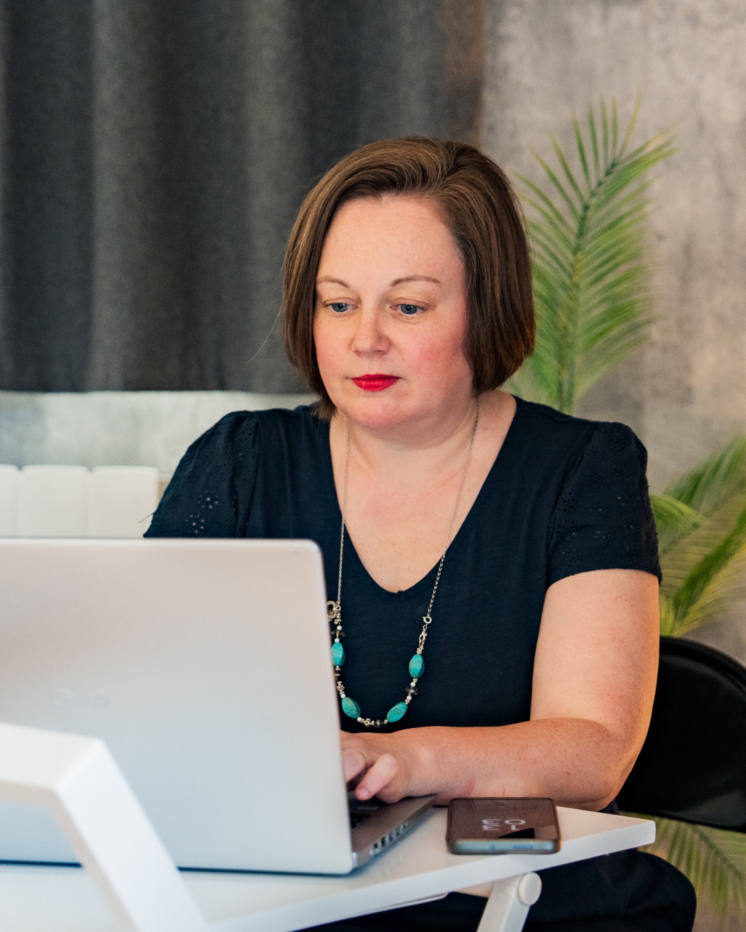 Woman with short brown hair working on a laptop at a desk, with a potted plant in the background.