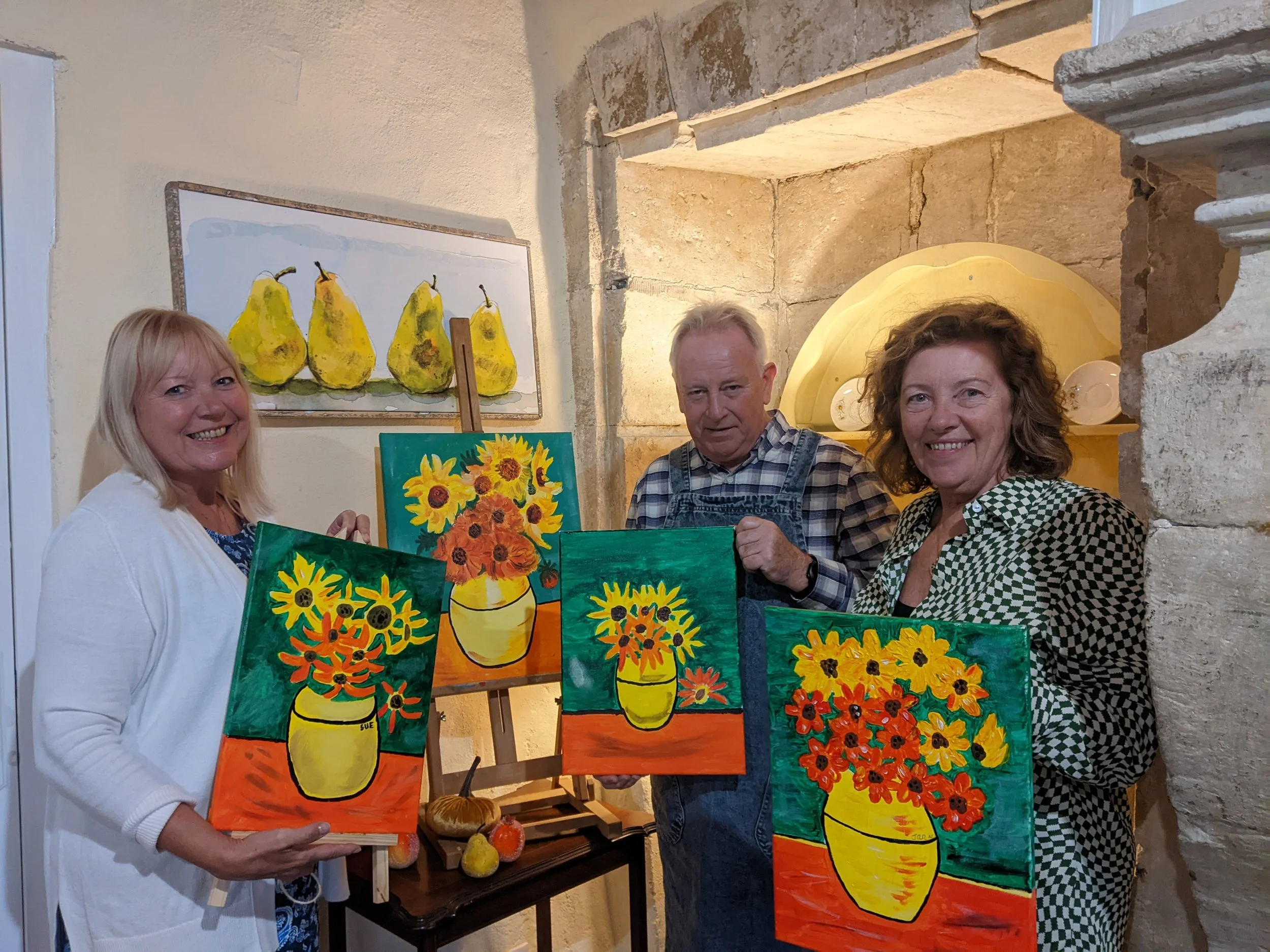 Three people standing indoors, holding paintings of sunflowers in yellow vases, with a backdrop of pear paintings on the wall.