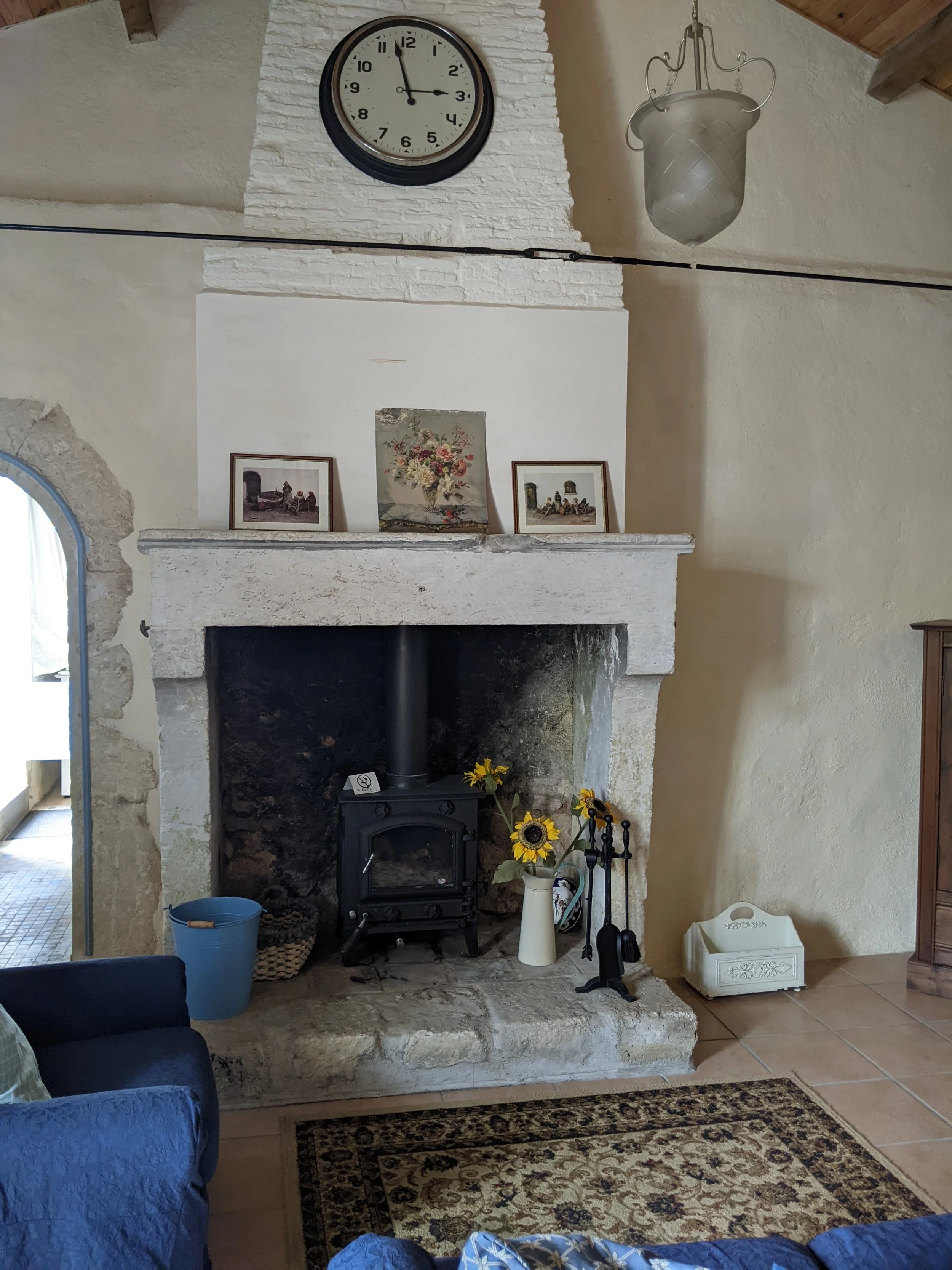 Living room with a stone fireplace and a black wood stove, decorated with sunflower arrangements in a white pitcher, framed pictures on the mantel, a wall clock, and a hanging light fixture.