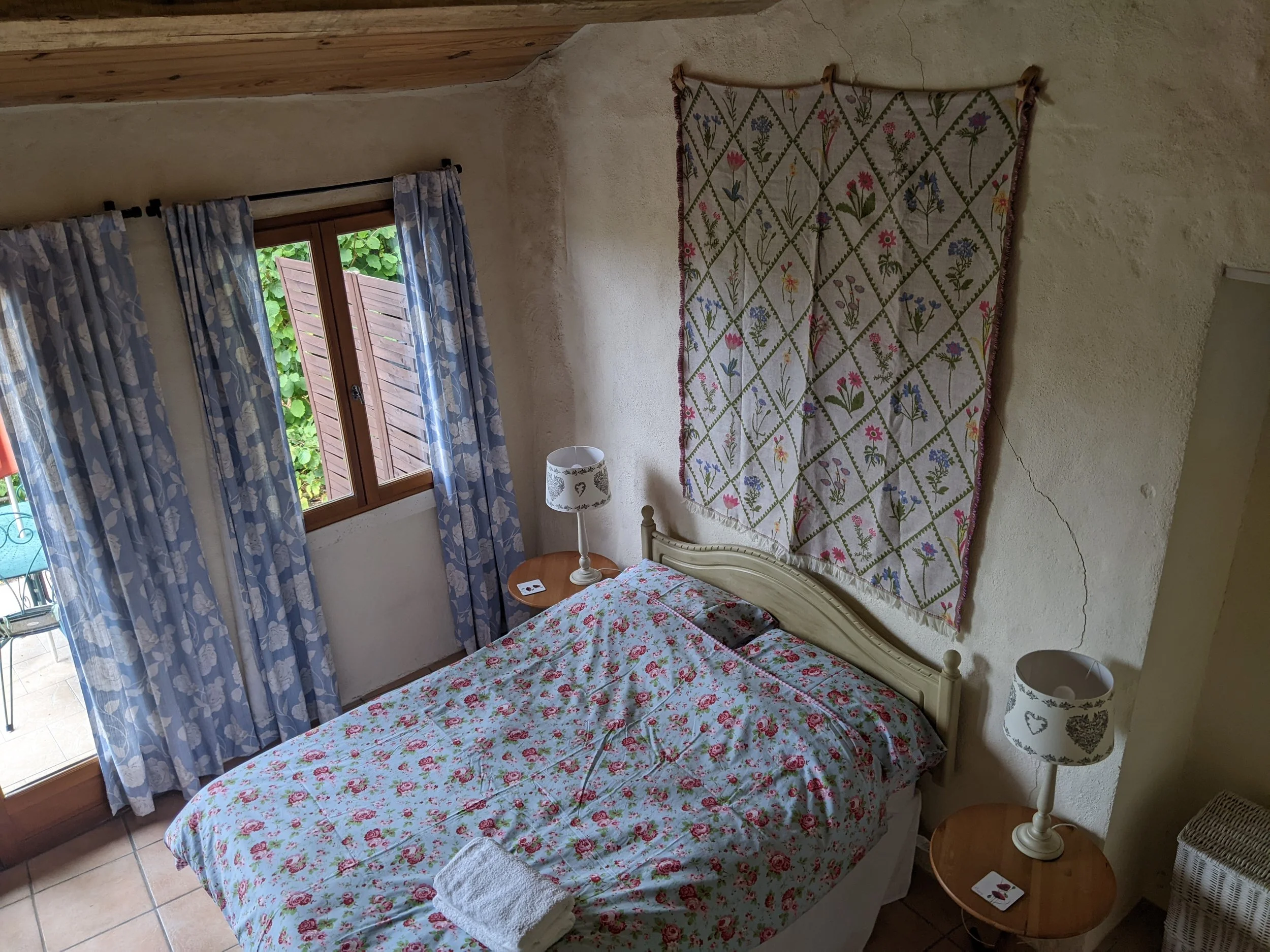 A cozy bedroom with a double bed covered in floral bedding, carved wooden headboard, two side tables with white lamps, a window with blue floral curtains, and a decorative floral tapestry on the wall.
