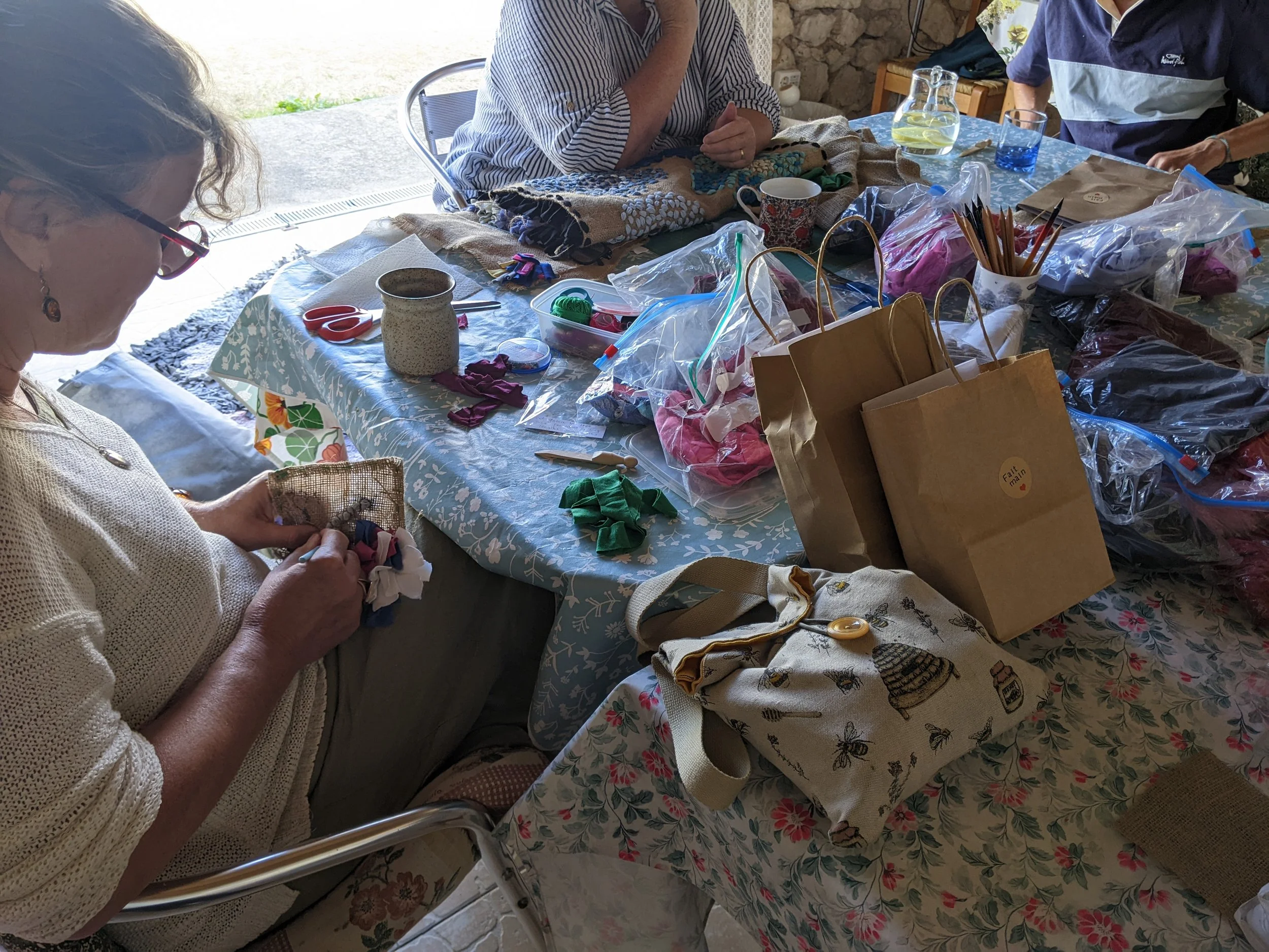 A woman sitting at a table engaged in fabric craft work, surrounded by various craft supplies, paper bags, and plastic storage bags filled with colorful materials, with other people also working at the same table.