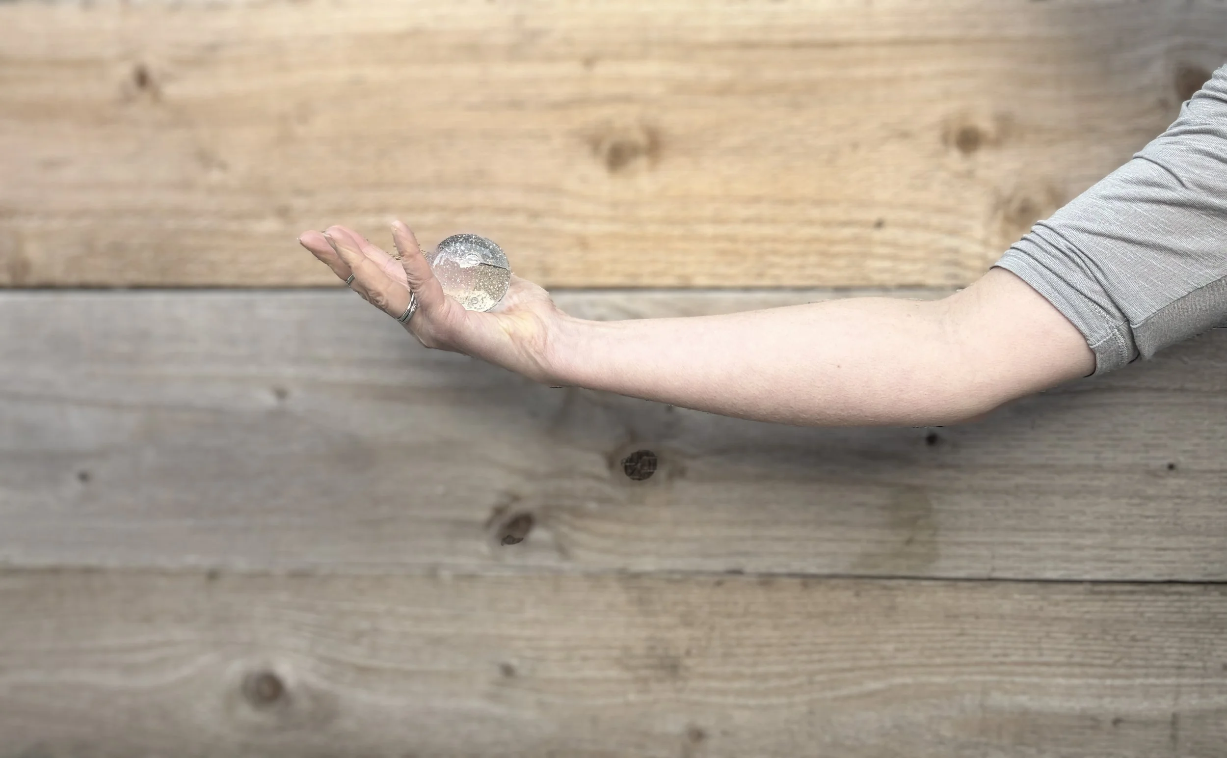 A person with a long arm and light skin holds a clear, round glass marble on their palm, against a wooden background.