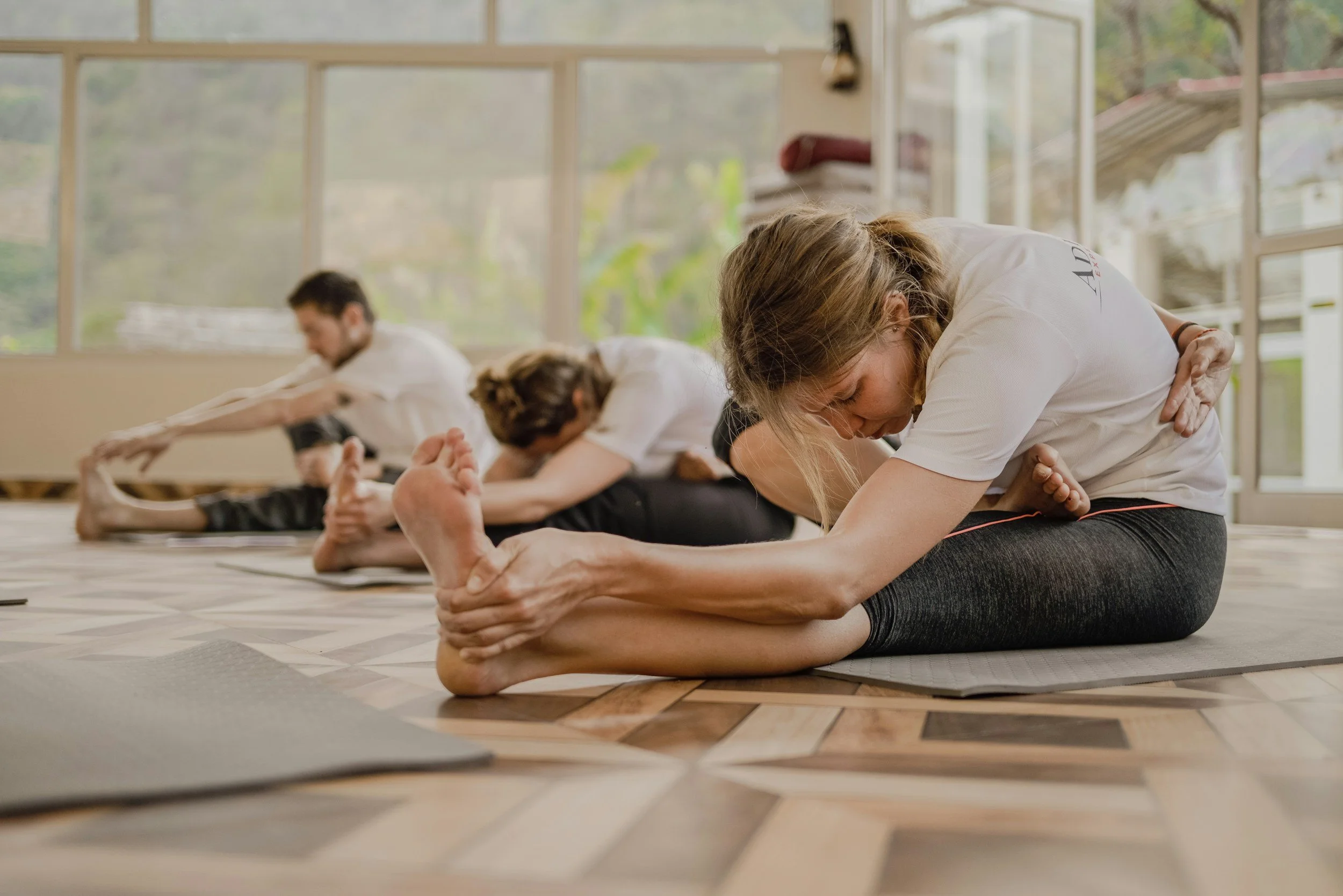 People practicing yoga in a bright, airy studio near large windows, sitting on yoga mats and stretching.