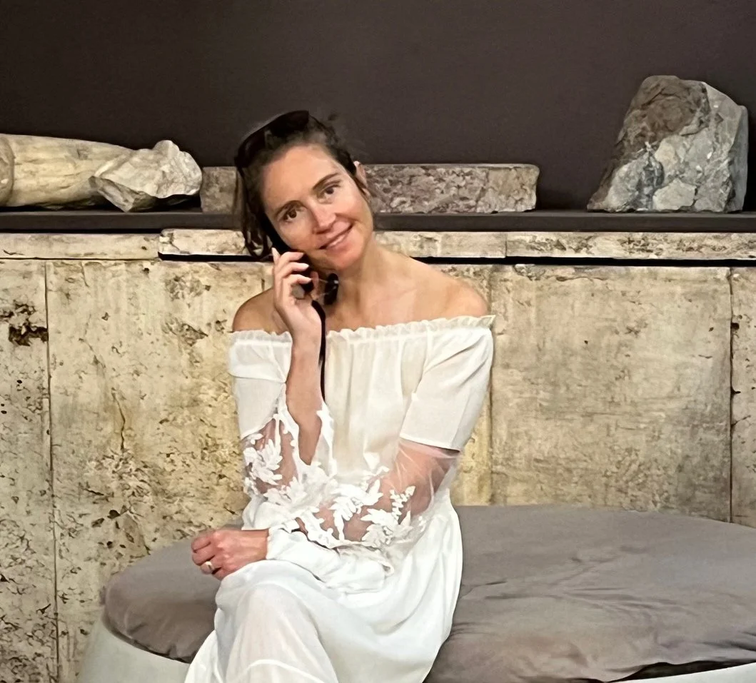 A woman with dark hair, wearing a white off-shoulder dress with lace sleeves, sitting on a cushioned surface, talking on a cellphone, with a stone wall and rocks in the background.