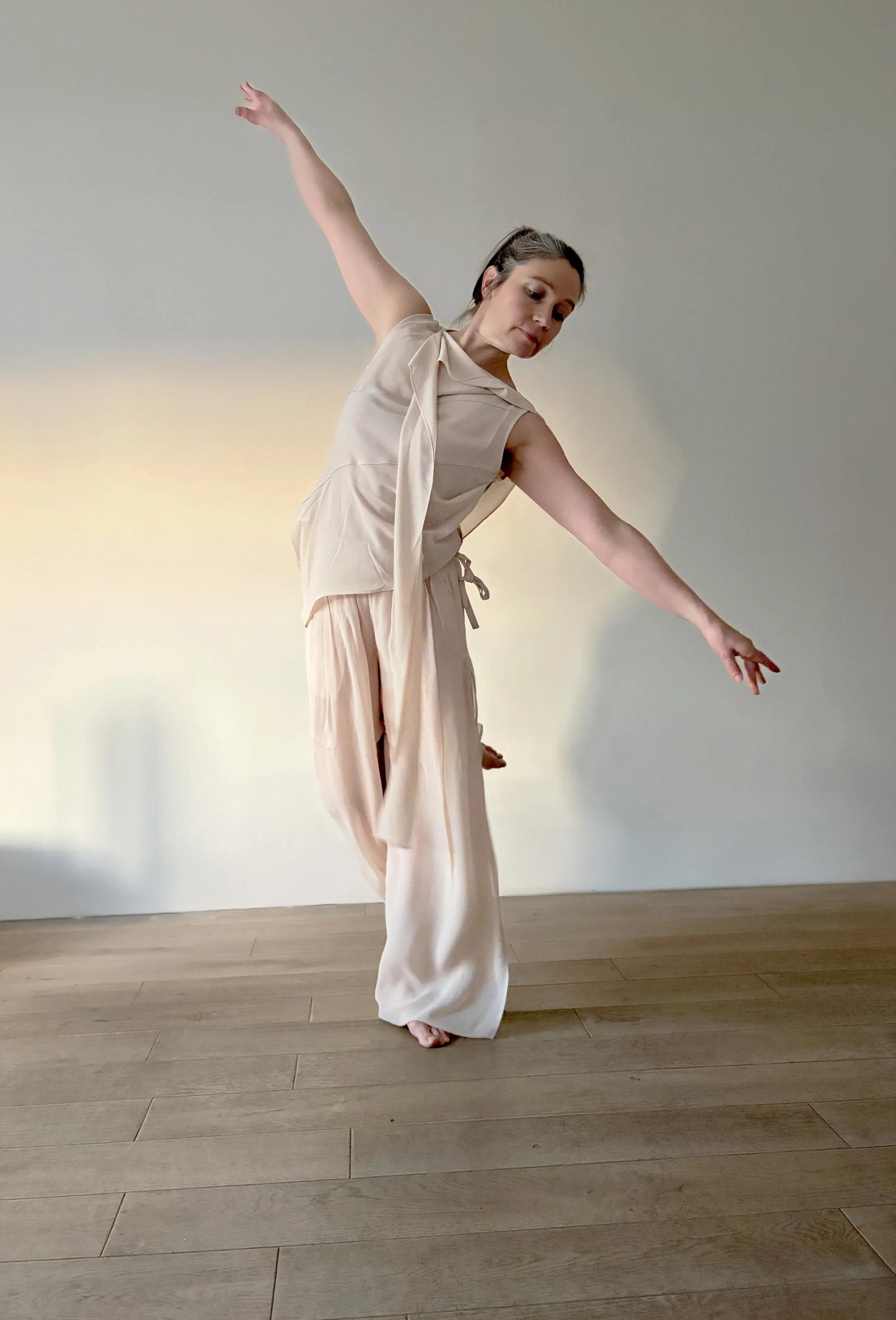 A woman in soft floaty clothing poses with arms extended in a dance pose on a wooden floor against a plain light-colored wall.
