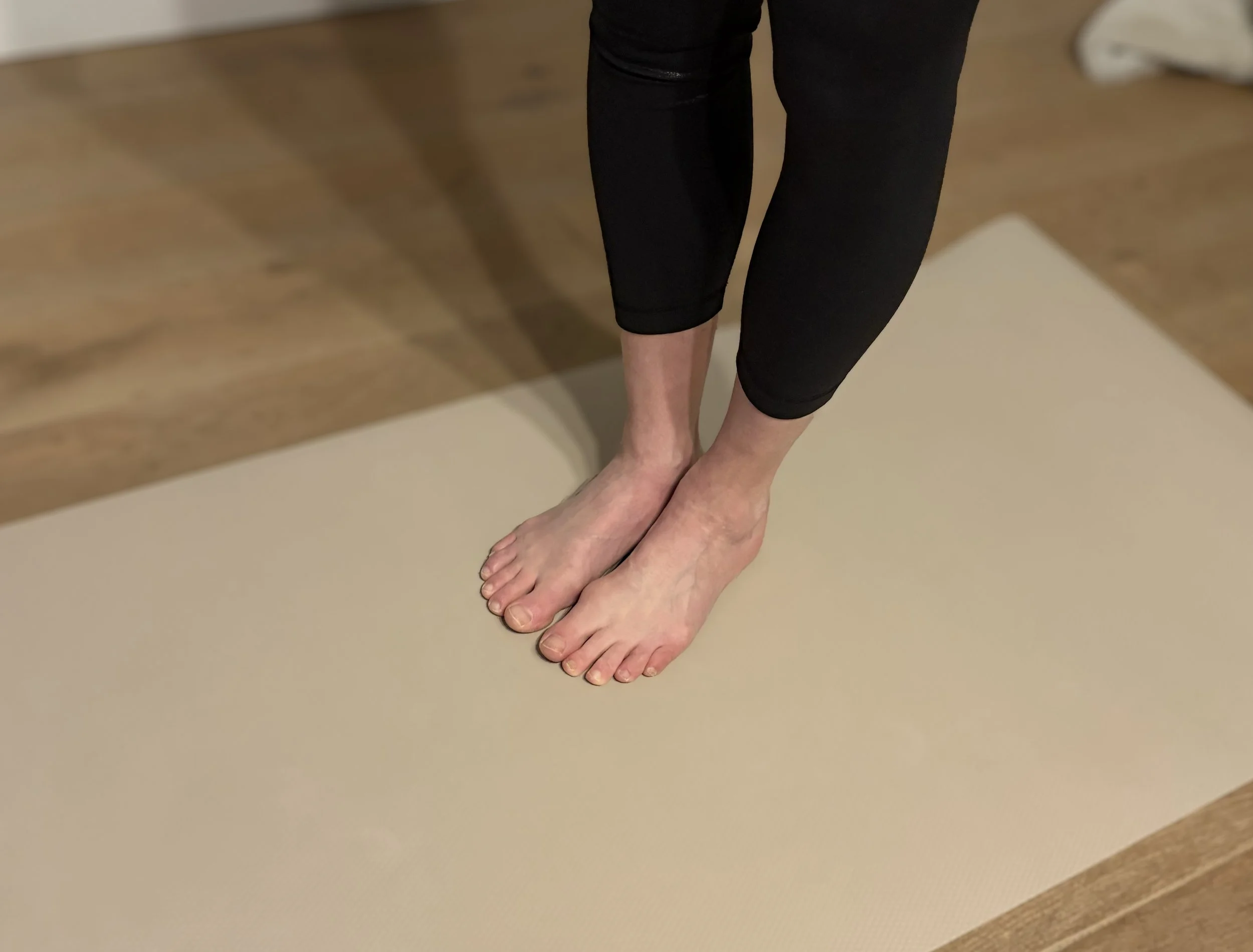 Person standing on a beige yoga mat in a room, wearing black leggings.