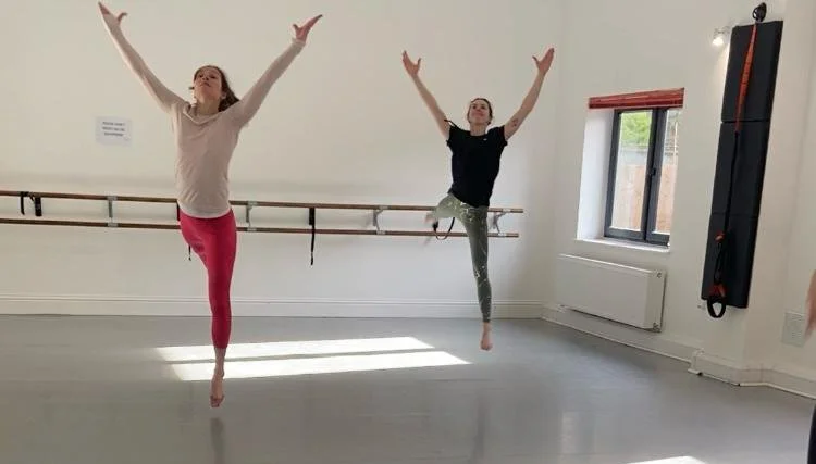 Two women practicing ballet or dance in a studio, balancing on one foot with arms raised, in front of a wall with a mirror and barre, near a window letting in natural light.