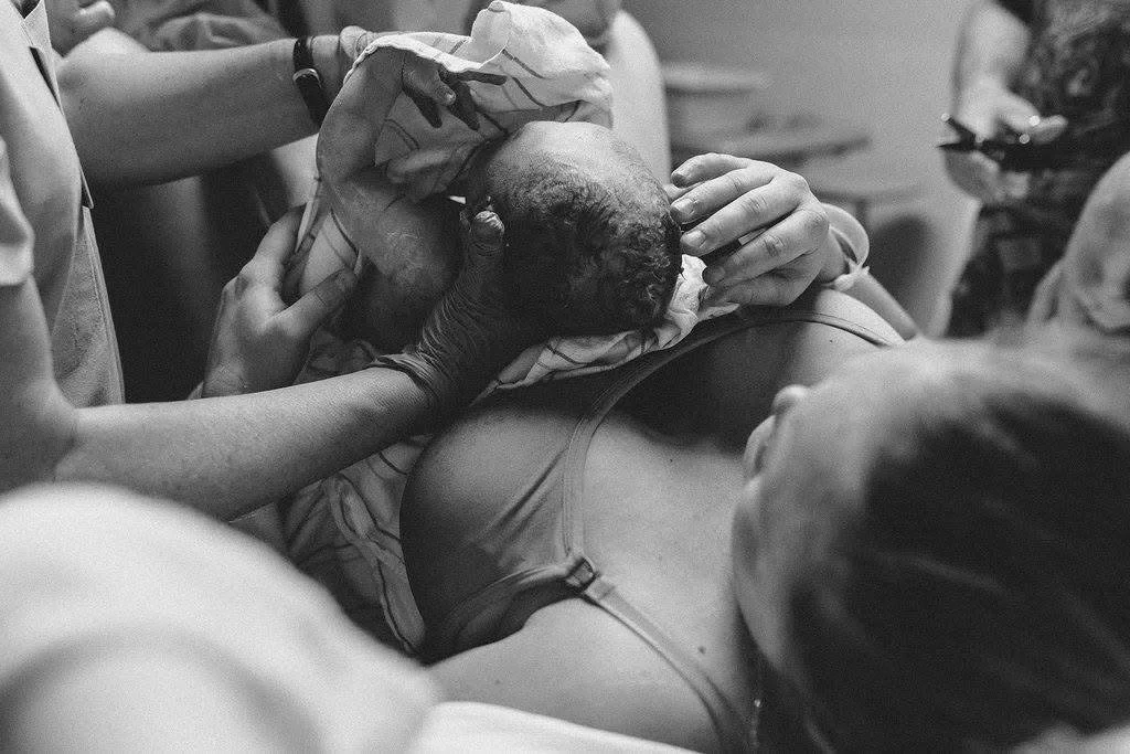 A woman giving birth in a hospital with medical staff assisting, holding her leg and supporting the baby's head as the baby is delivered.