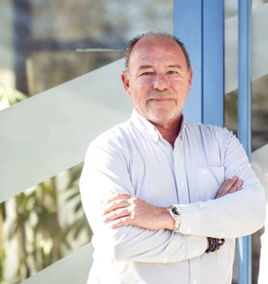 Homme d'âge moyen avec une chemise blanche, portant une montre et des bracelets, croisant les bras, posant devant une fenêtre moderne.