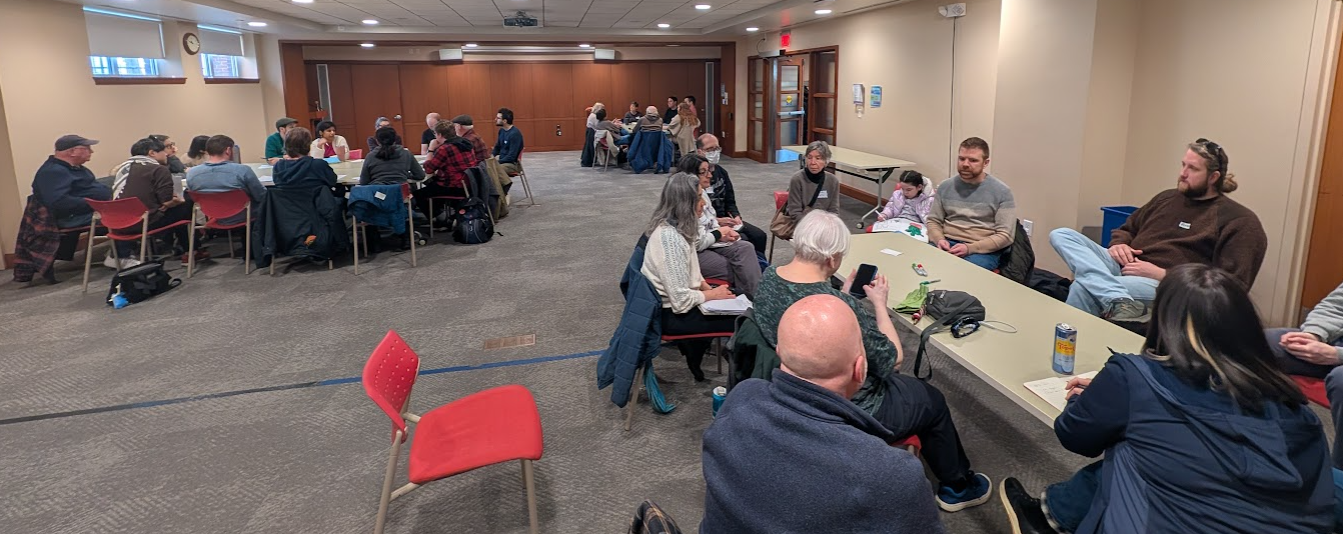 People are seated in three different groups in a large meeting room at the main library