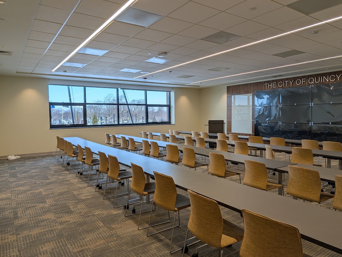 Meeting room with four rows of tables, each with 12 chairs. There are screens at the front of the room and large windows on the side of the room. This is a community room at the new public safety building on Sea Street