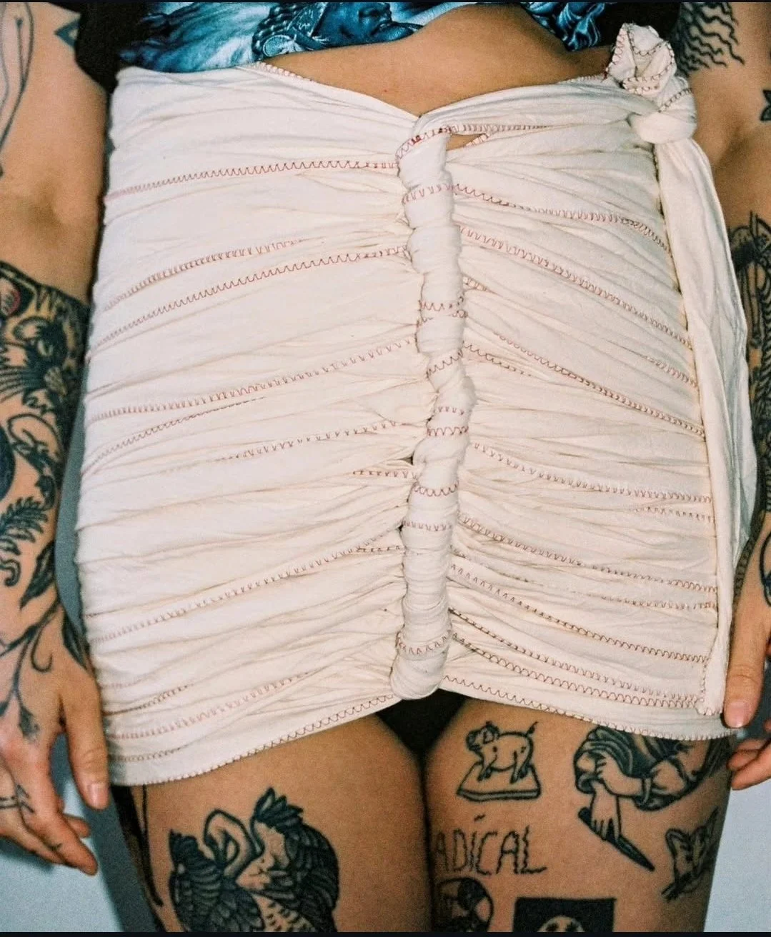 De foto toont een persoon met getatoeëerde armen en benen, die een wit gebleekte doek met opvallende stiksels en rimpelingen vasthoudt.