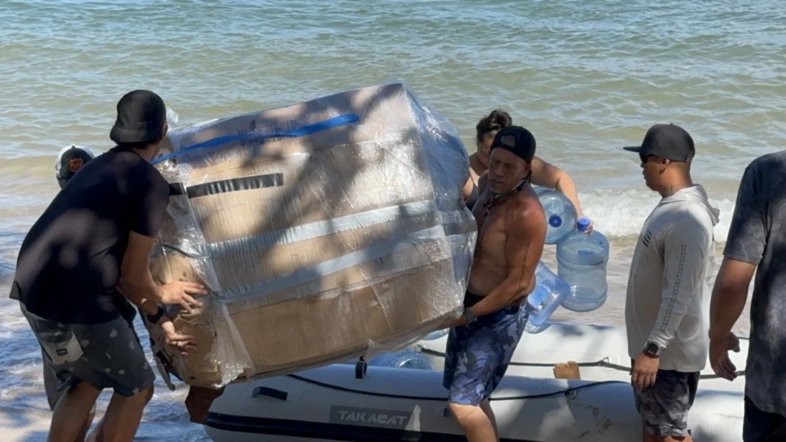 Group of people transporting a large, wrapped package from a small boat onto a beach.