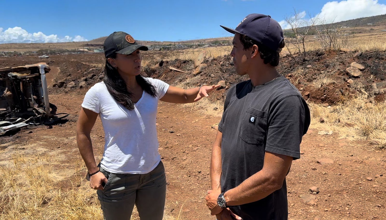 Two people standing in a dry, barren landscape with an overturned vehicle in the background.