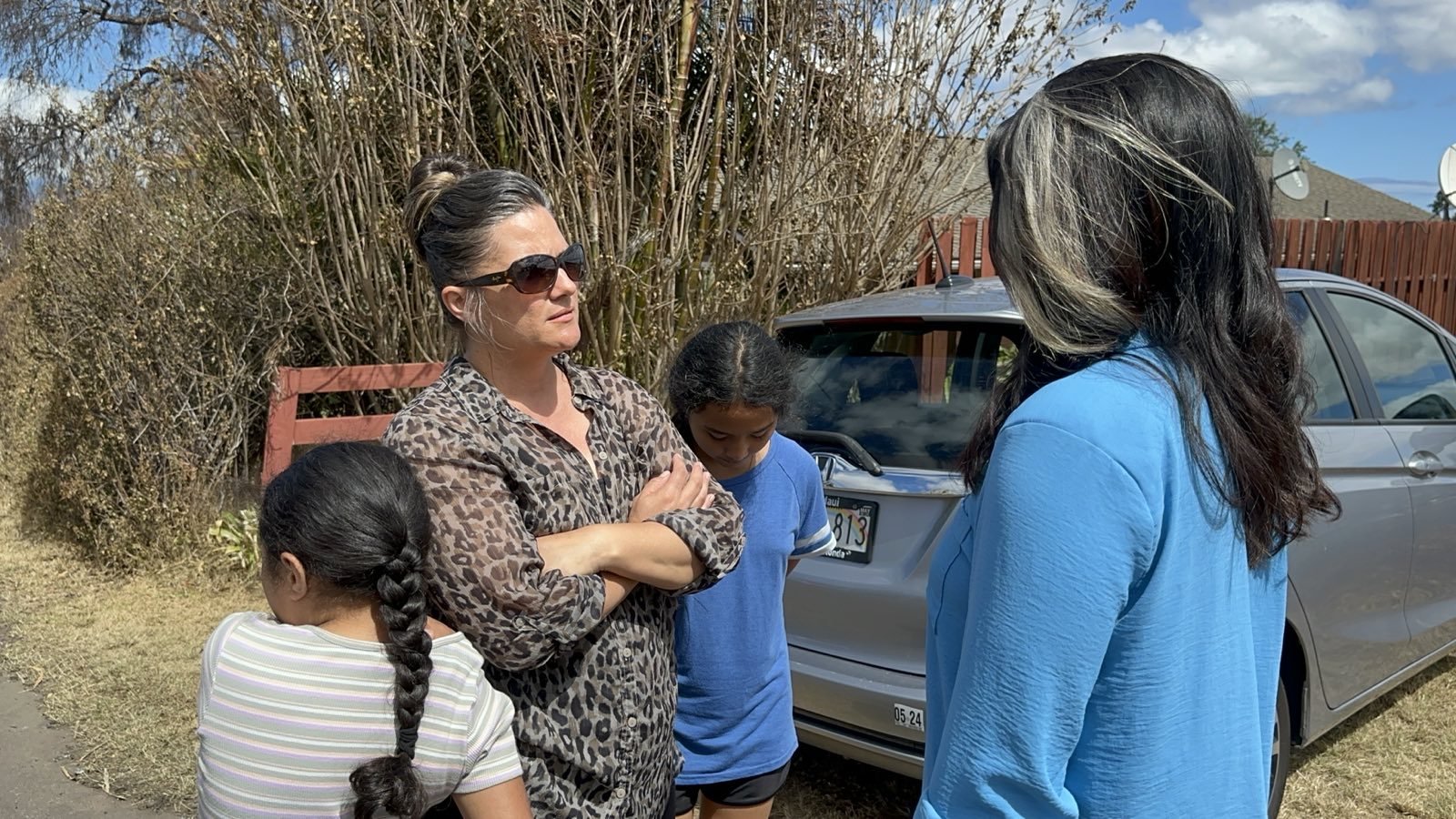A woman wearing sunglasses and a patterned shirt stands with her arms crossed, speaking to another woman with long hair wearing a blue shirt. Two children stand nearby, with one child hugging the woman's waist. A car is parked in the background next