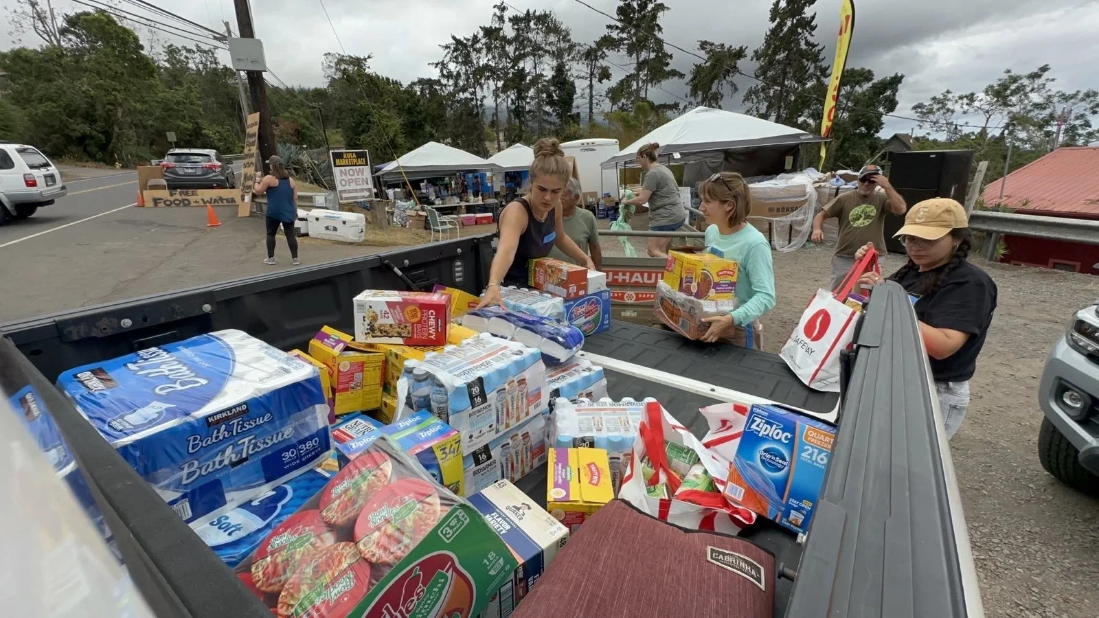 People loading supplies into a pickup truck, including bottled water, paper towels, and food items, at a roadside relief station with signs for free food and water.