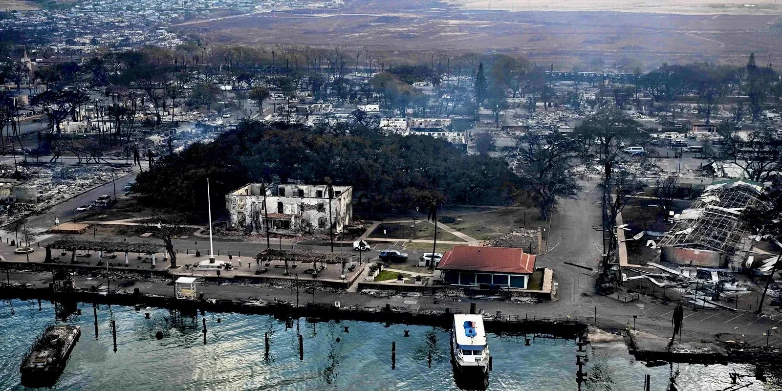 Aerial view of a coastal area with fire-damaged buildings and debris. Trees and structures are charred, with visible destruction across the landscape. A boat is docked near the waterfront.