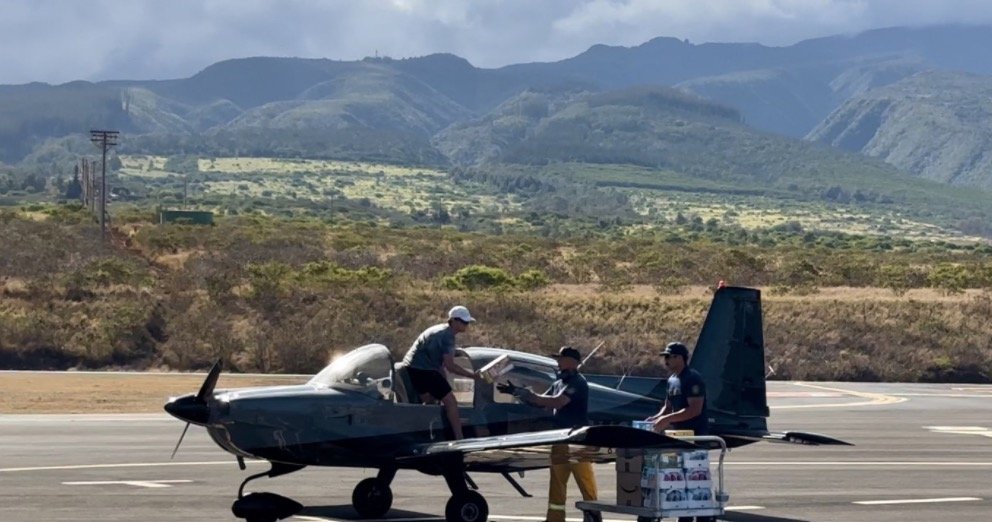 Small aircraft on runway with three people near it and mountainous landscape in the background.