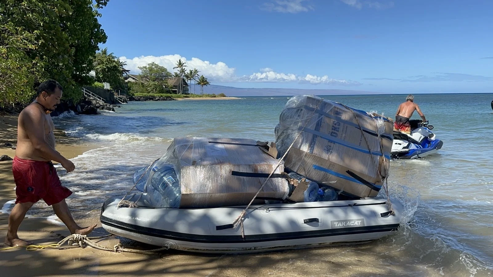 Man pulling an inflatable boat loaded with large wrapped packages and water bottles on a beach, another person on a jet ski in the water.