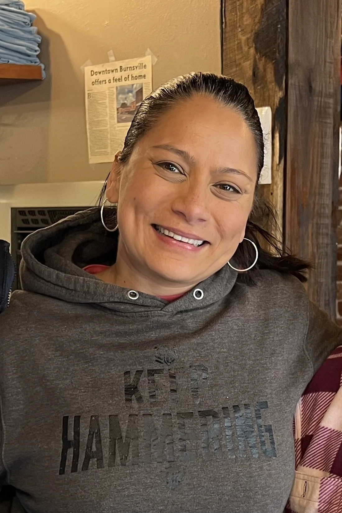 A smiling person wearing a gray hoodie with text "KEEP HAMMERING" printed on it, standing indoors near a wooden wall. A newspaper clipping is visible in the background.
