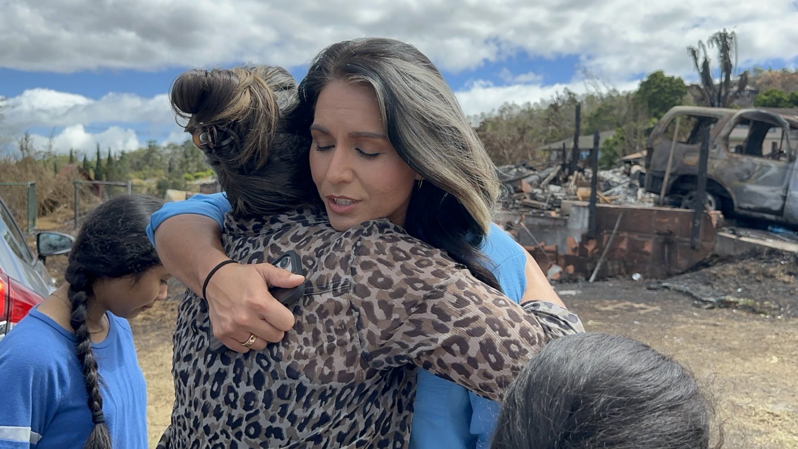 A woman in a leopard print top hugs another woman with long dark hair. They are standing outdoors near burnt debris and a charred vehicle under a partly cloudy sky. A person with a braid is partially visible on the left.