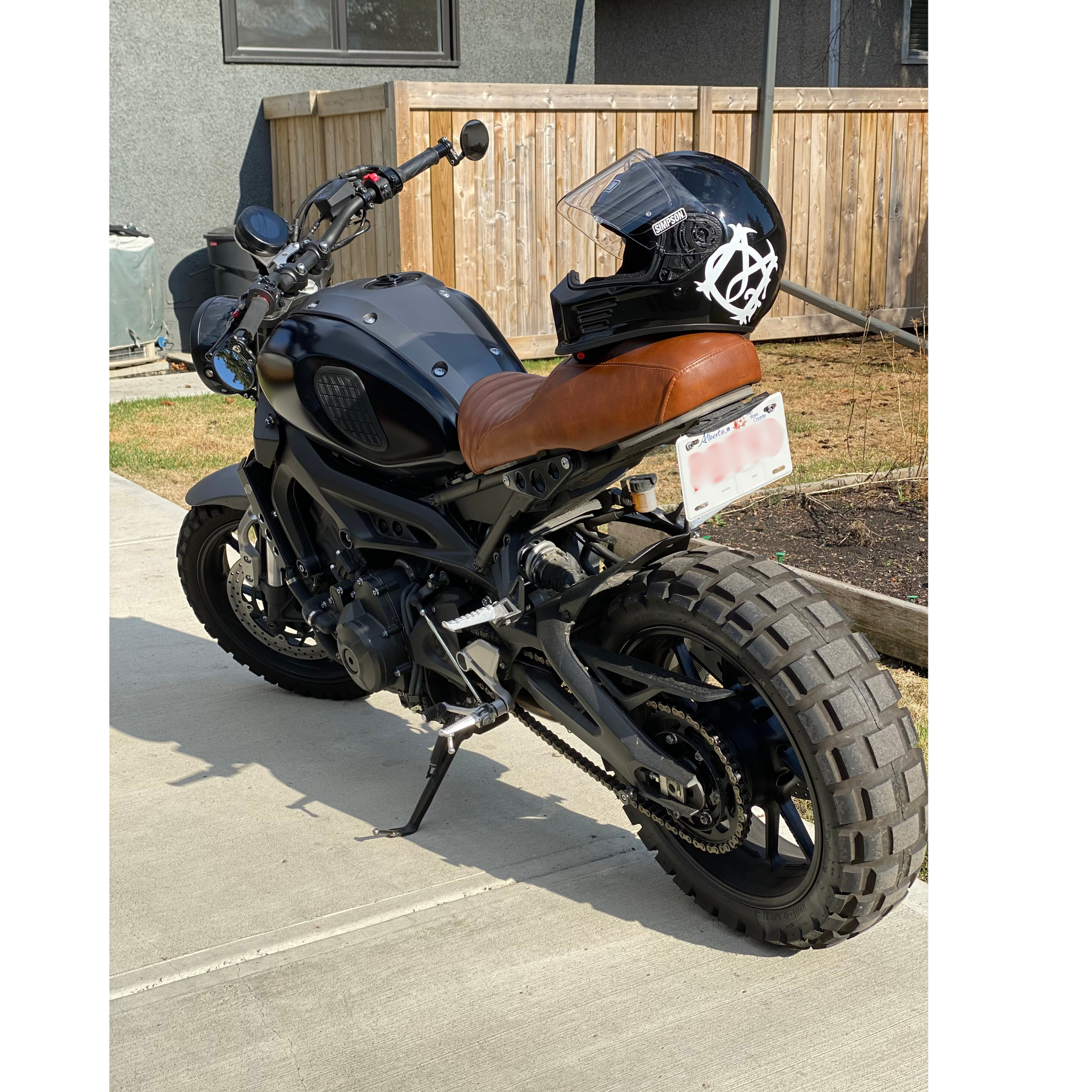A black motorcycle with a brown leather seat parked on a concrete driveway, with a black helmet resting on the seat. The helmet has a white graphic design. There is a wooden fence and part of a house in the background.