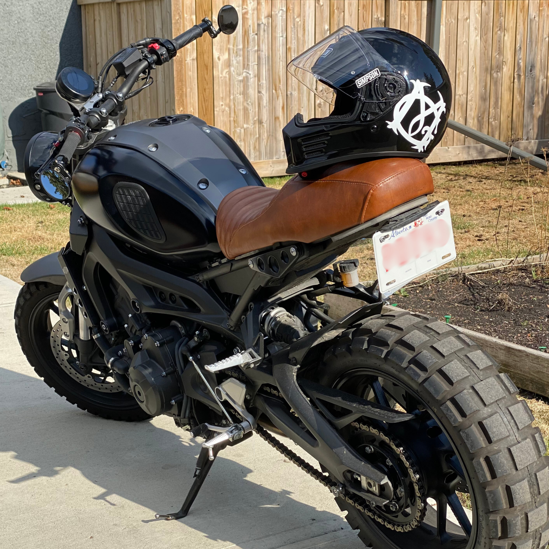A black motorcycle with a brown leather seat and a black full-face helmet resting on the seat. The motorcycle is parked on a concrete driveway in front of a wooden fence. The motorcycle has large off-road tires, a digital dashboard, and side mirrors. The license plate is partially visible, showing the state of Alberta.