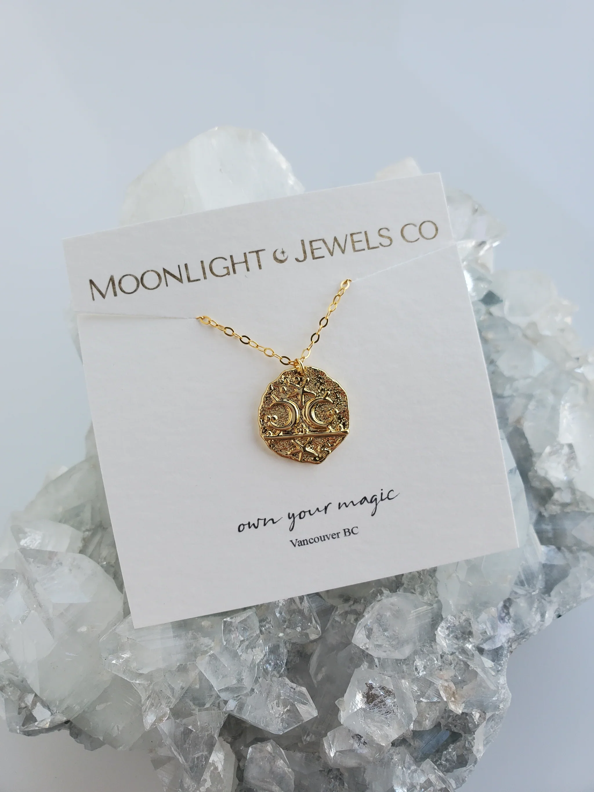 Gold necklace with a circular pendant featuring a decorative design, displayed on a card that reads 'MOONLIGHT & JEWELS CO' and 'own your magic' from Vancouver BC, placed on a bed of clear crystal rocks.
