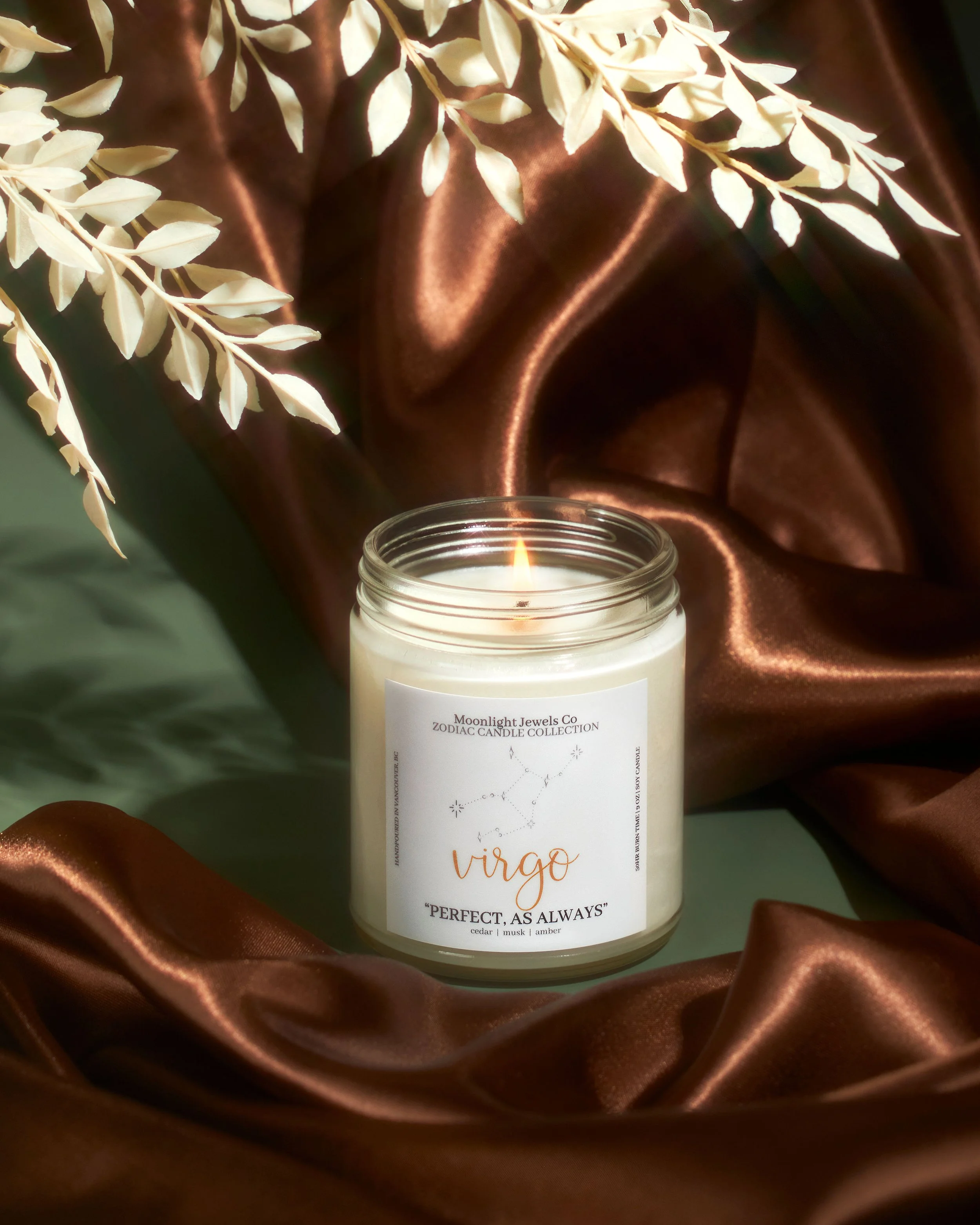 A lit candle in a glass jar with a Virgo zodiac sign label, surrounded by brown satin fabric and white dried leaves.