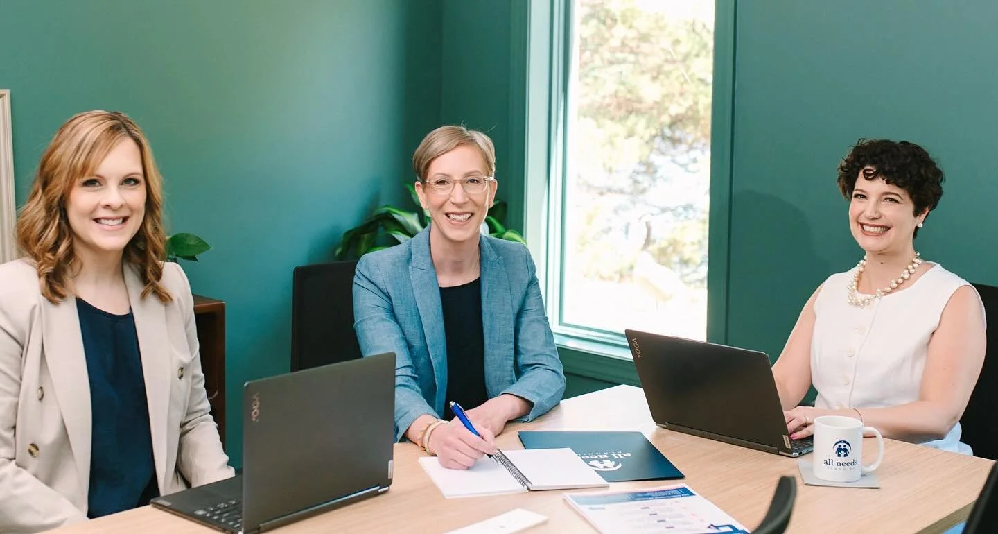 Three women sitting at a conference table with laptops and documents in a meeting room with green walls and a window.