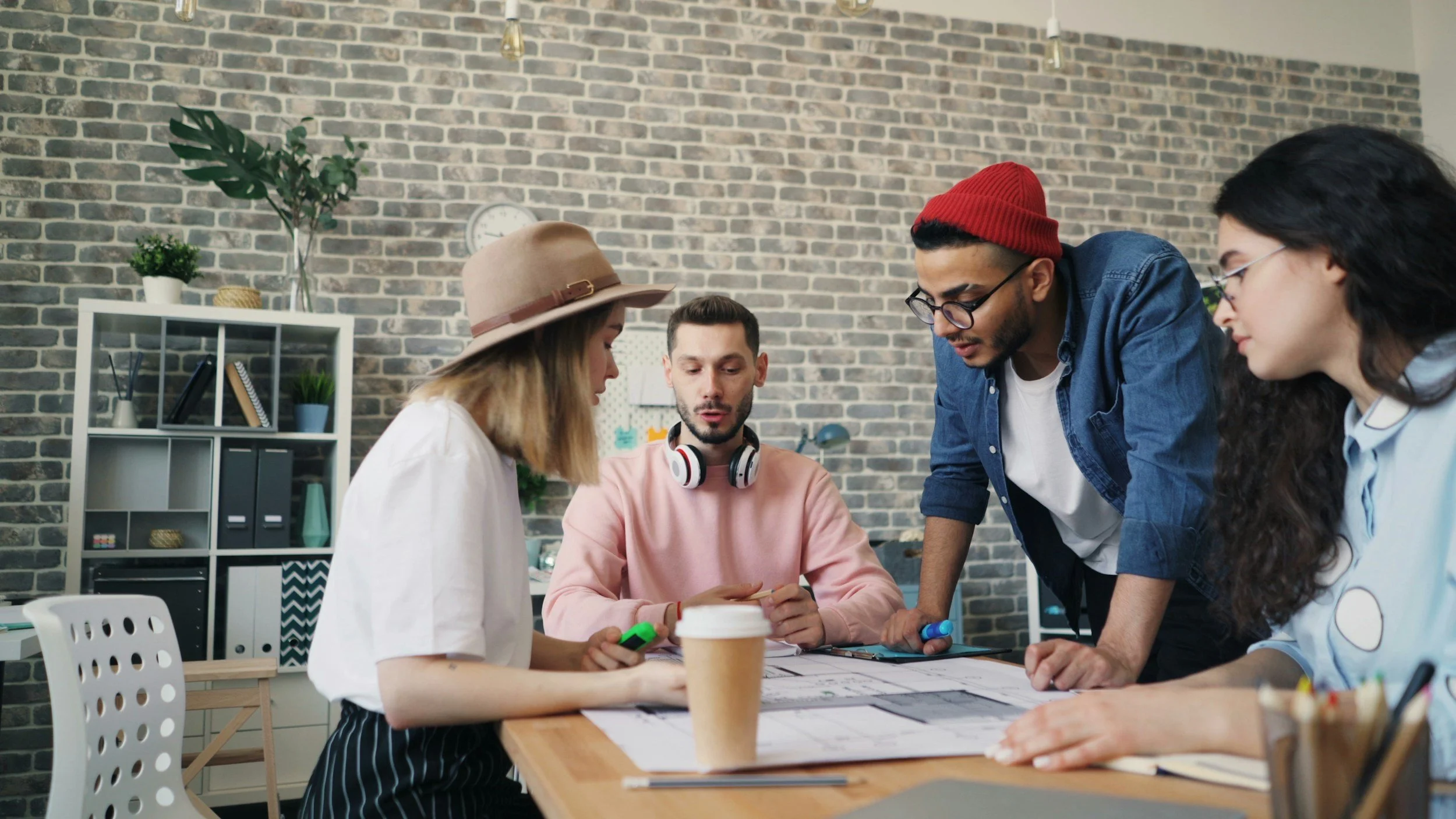 Four young adults in a meeting room with a brick wall background, gathered around a table with paper plans, a coffee cup, and a tablet, discussing a project.