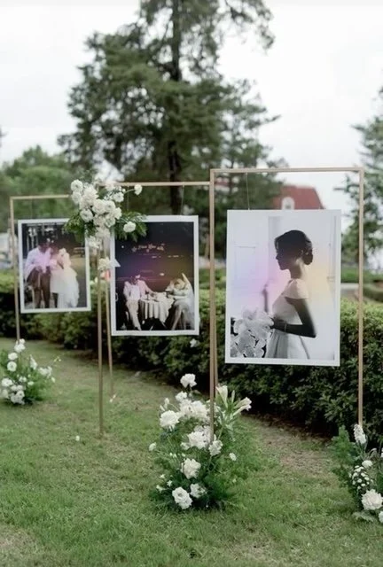 Outdoor display of large framed photographs decorated with white flowers on a grassy area, with trees and a building visible in the background.