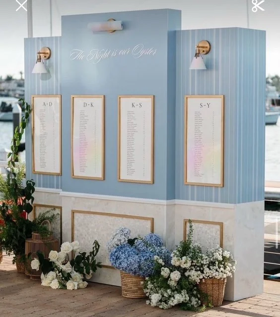 A wedding seating chart display with four framed sections labeled A-D, D-K, K-S, and S-Y, mounted on a blue and white wall. Decorative flowers in baskets and vases are placed at the base of the display.