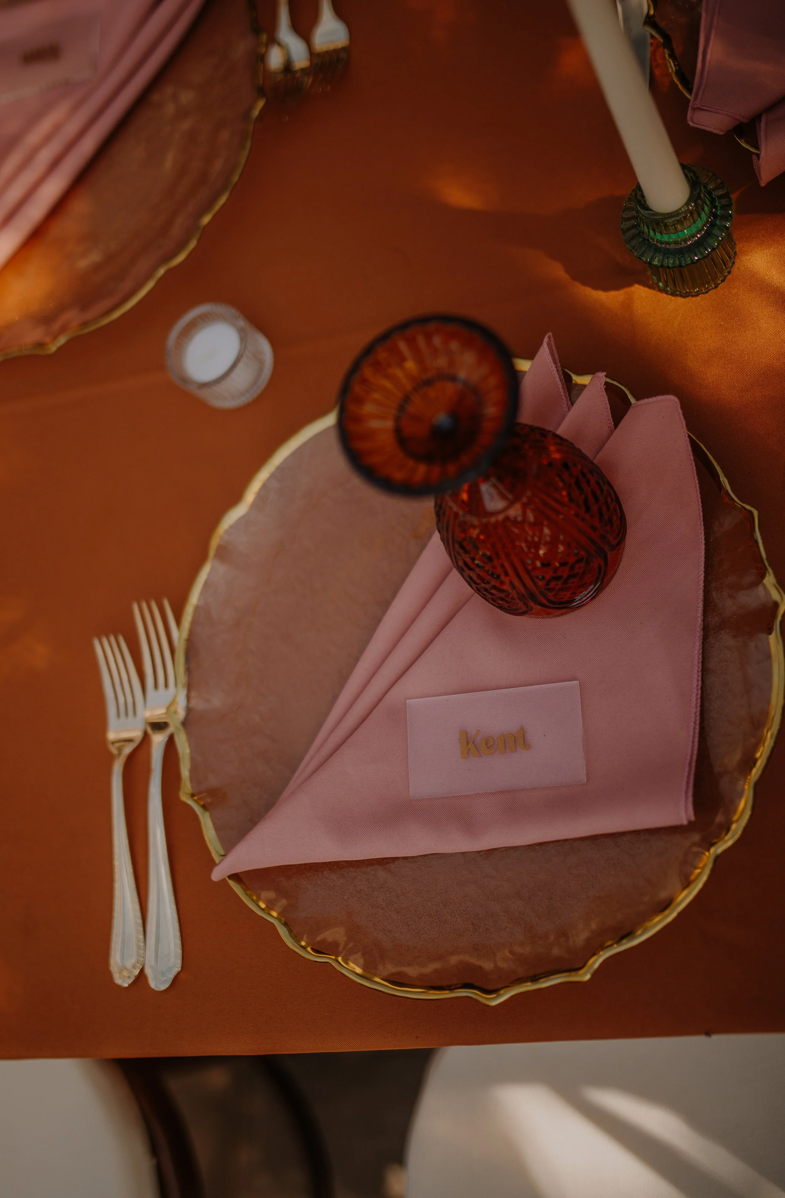 Elegant table setting with a pink napkin labeled 'Kent', a green glass holder, a red glass vase, two forks, a brass-edged pink tablecloth, and decorative is:ay and tableware.
