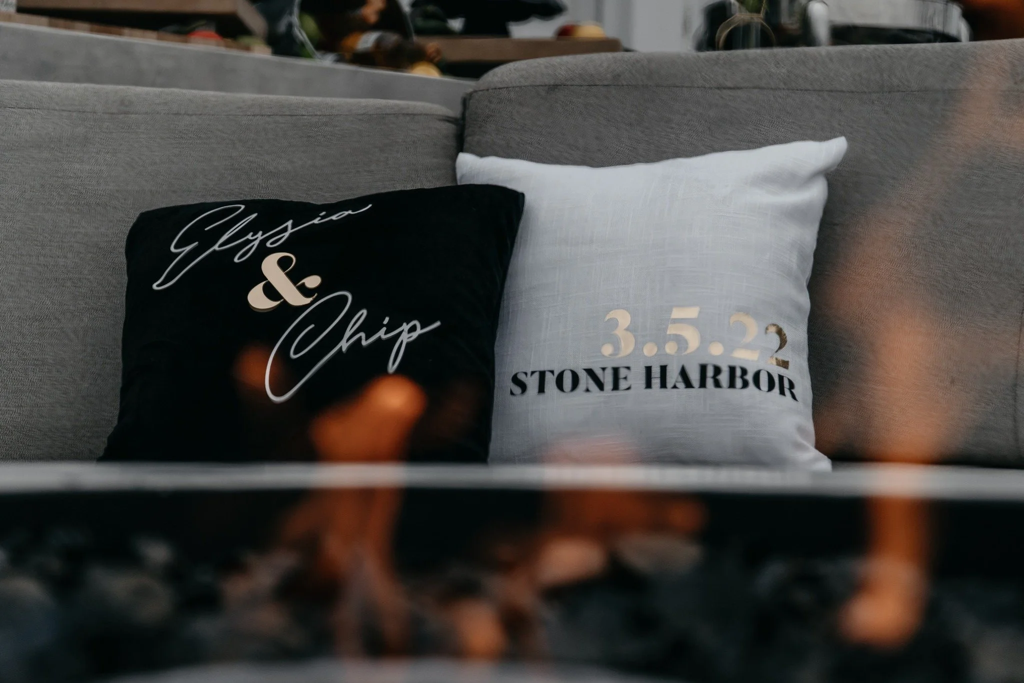 Two decorative pillows on a gray couch, one black with white cursive text reading 'Glysia & Chip,' and one white with black text displaying the date '3.5.22' and the location 'Stone Harbor.'