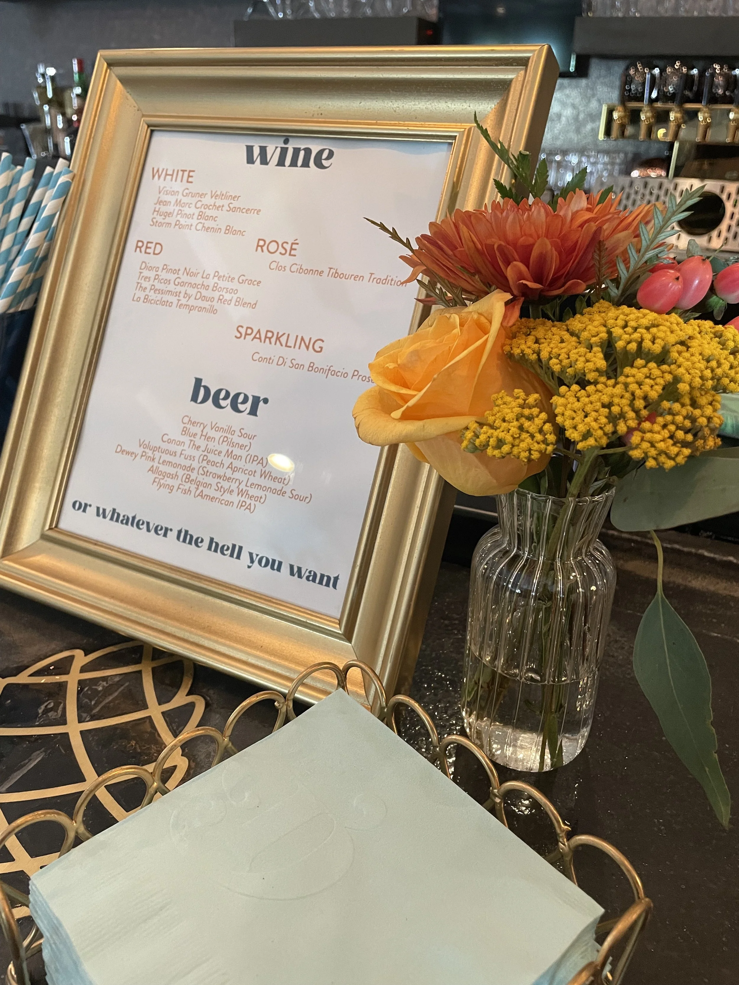 A menu board listing various wines and beers, displayed in a gold frame, next to a vase of colorful flowers on a table at a bar or restaurant.